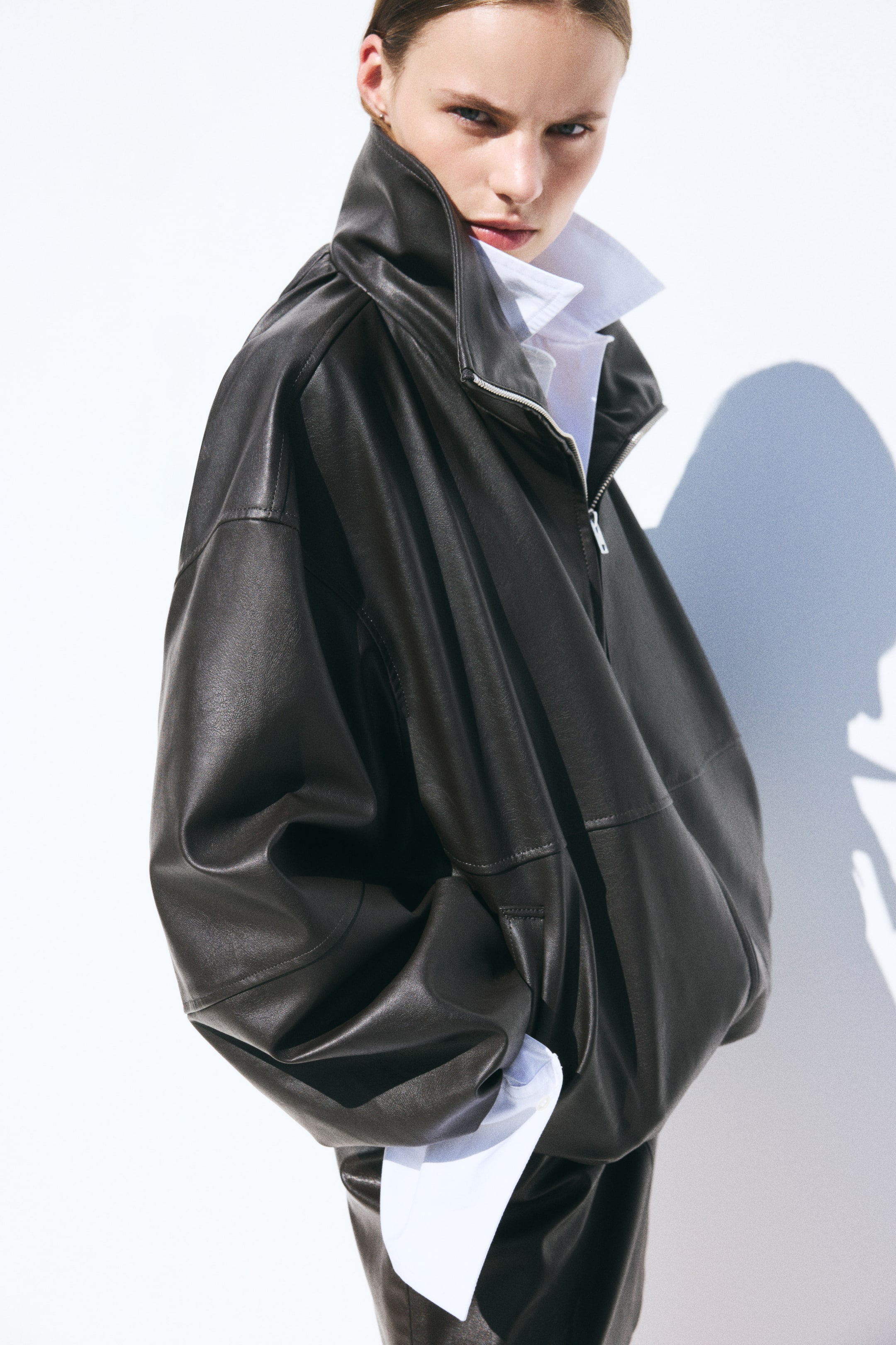 View larger image: The model is wearing an oversized jacket in dark coated fabric, layered over a white collared shirt. The jacket features a high funnel collar, dropped shoulders, and a welt side pocket where the model is placing their hand.