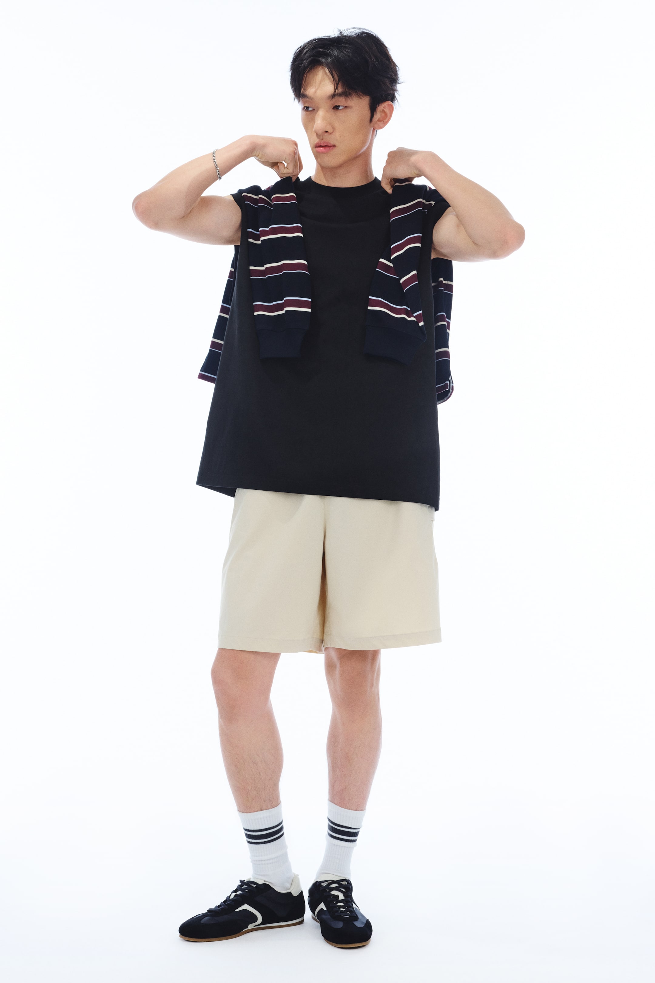 Xem ảnh lớn hơn: Một chiếc áo vest đen bằng vải cotton jersey dày với dáng rộng đang được người mẫu mặc. Áo có cổ tròn viền gân, lỗ tay áo có viền thô và gấu áo cắt thẳng; người mẫu đang chỉnh sửa cổ áo bằng cả hai tay trong khi một chiếc áo len sọc được khoác lên vai.