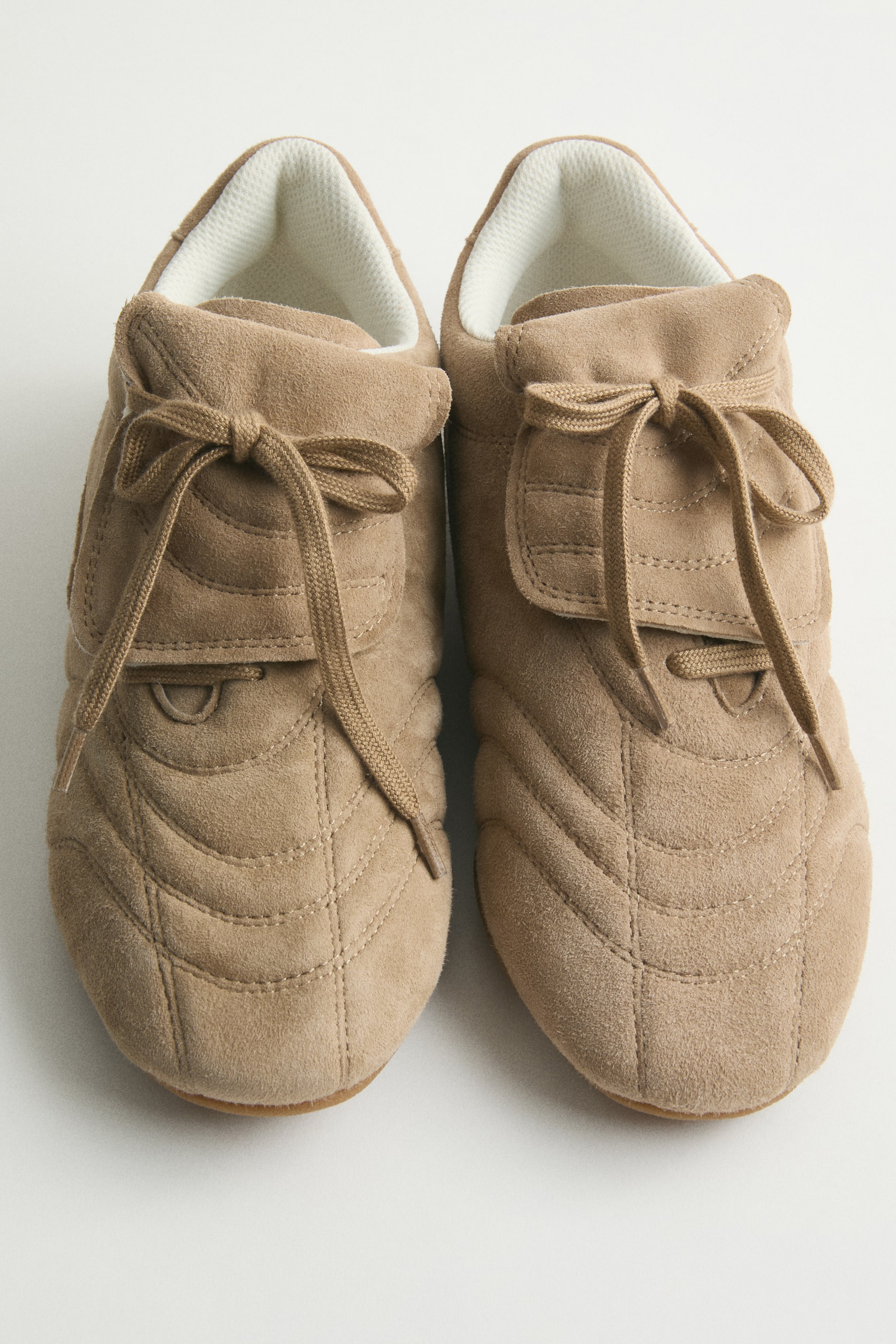 Agrandir l'image: Une paire de chaussures en daim beige présente un motif de surpiqûres matelassées sur l'empeigne et les côtés, avec des lacets assortis noués en nœud au-dessus de la languette, et une doublure en maille blanche est visible autour du col.