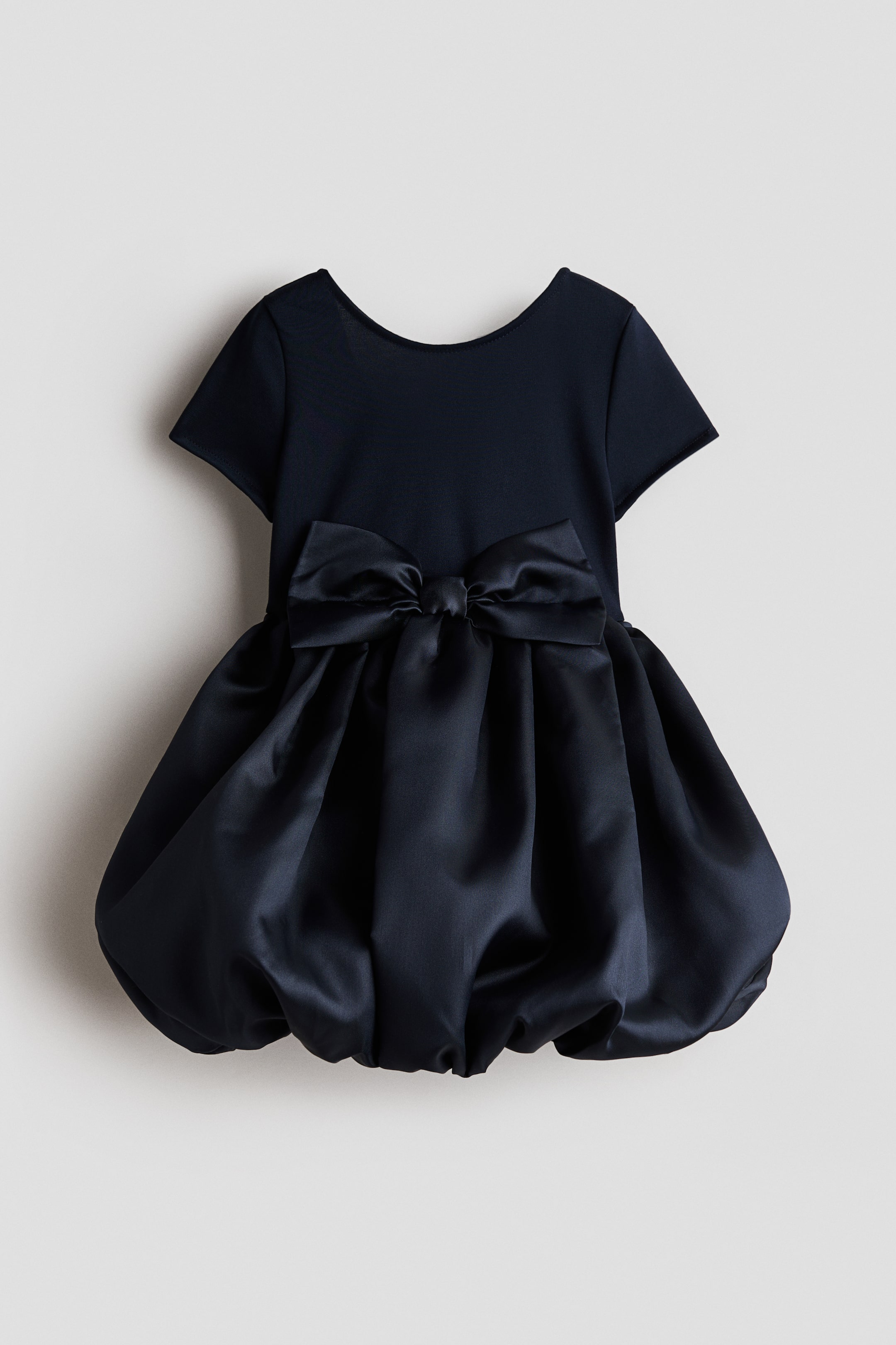 View larger image: A navy blue dress is showcasing its back view, featuring a matte knit bodice with short sleeves and a scoop neckline. A large satin bow embellishes the waist, flowing into a voluminous, gathered satin bubble skirt.