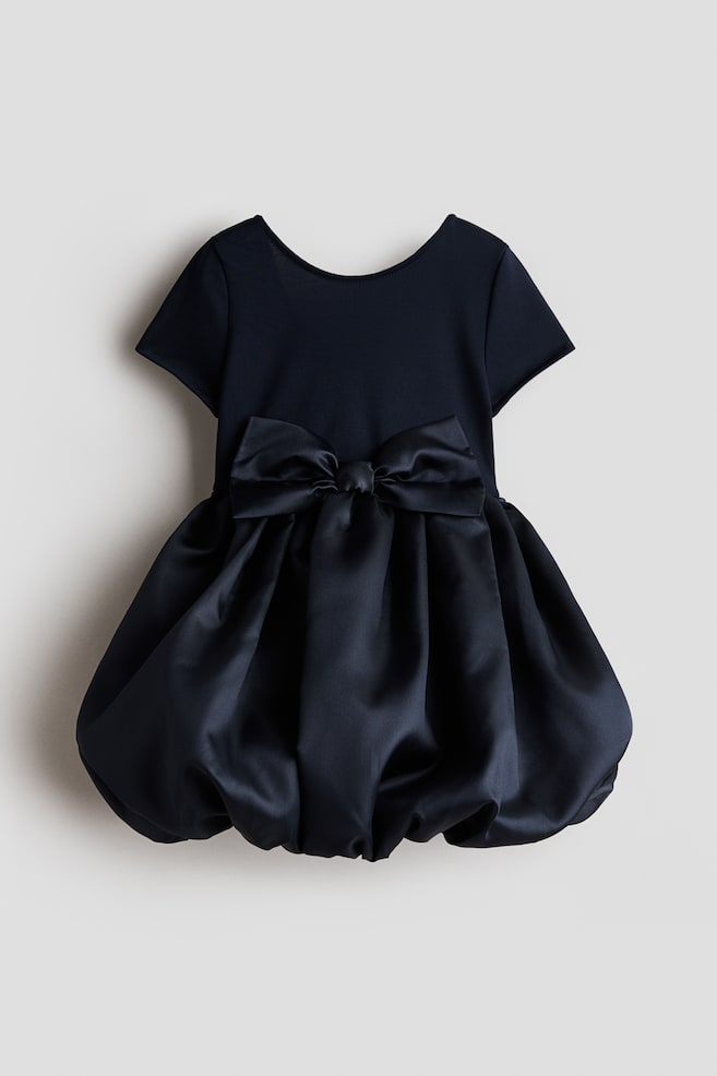 A navy blue dress is showcasing its back view, featuring a matte knit bodice with short sleeves and a scoop neckline. A large satin bow is embellishing the waist, which is flowing into a voluminous, gathered satin bubble skirt. - 1