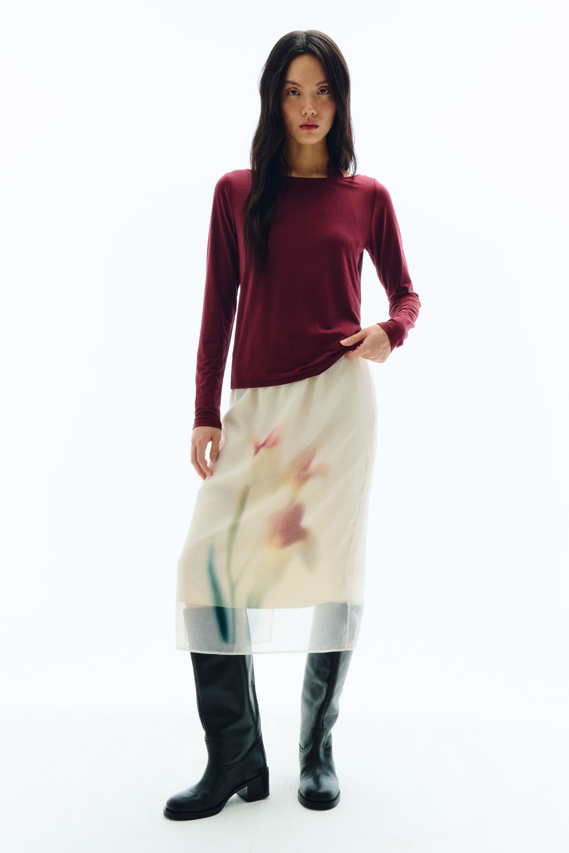 拡大画像を表示: ワイドなネックラインが特徴のマルーンカラーの長袖トップスが、シアーなオフホワイトのミディ丈スカートとコーディネートされています。スカートにはソフトなピンク、イエロー、グリーンで描かれた抽象的なフラワーパターンがあしらわれ、ブラックのニーハイブーツの上にレイヤードされています。