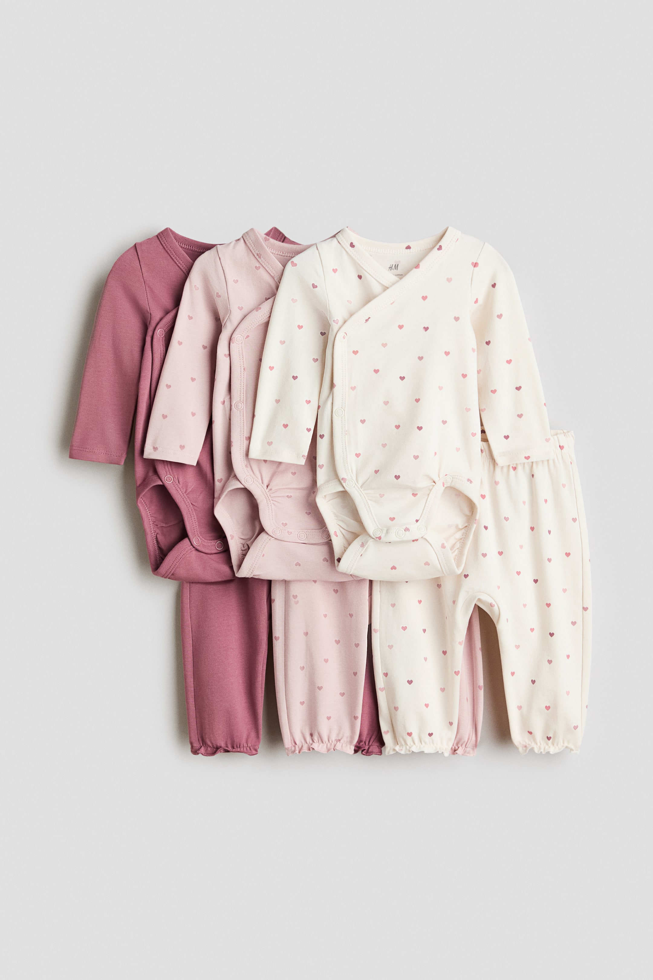 View larger image: Presenting a set of three baby bodysuits and three pairs of trousers, artfully arranged from left to right in delightful shades of pink and cream. The long-sleeved bodysuits feature wrapover fronts with side press-studs and gathered leg seams, ensuring both style and comfort. Complementing them, the matching trousers showcase wide foldover waist panels and cuffed hems, with two sets adorned with a charming heart pattern. Perfect for your little one's wardrobe!