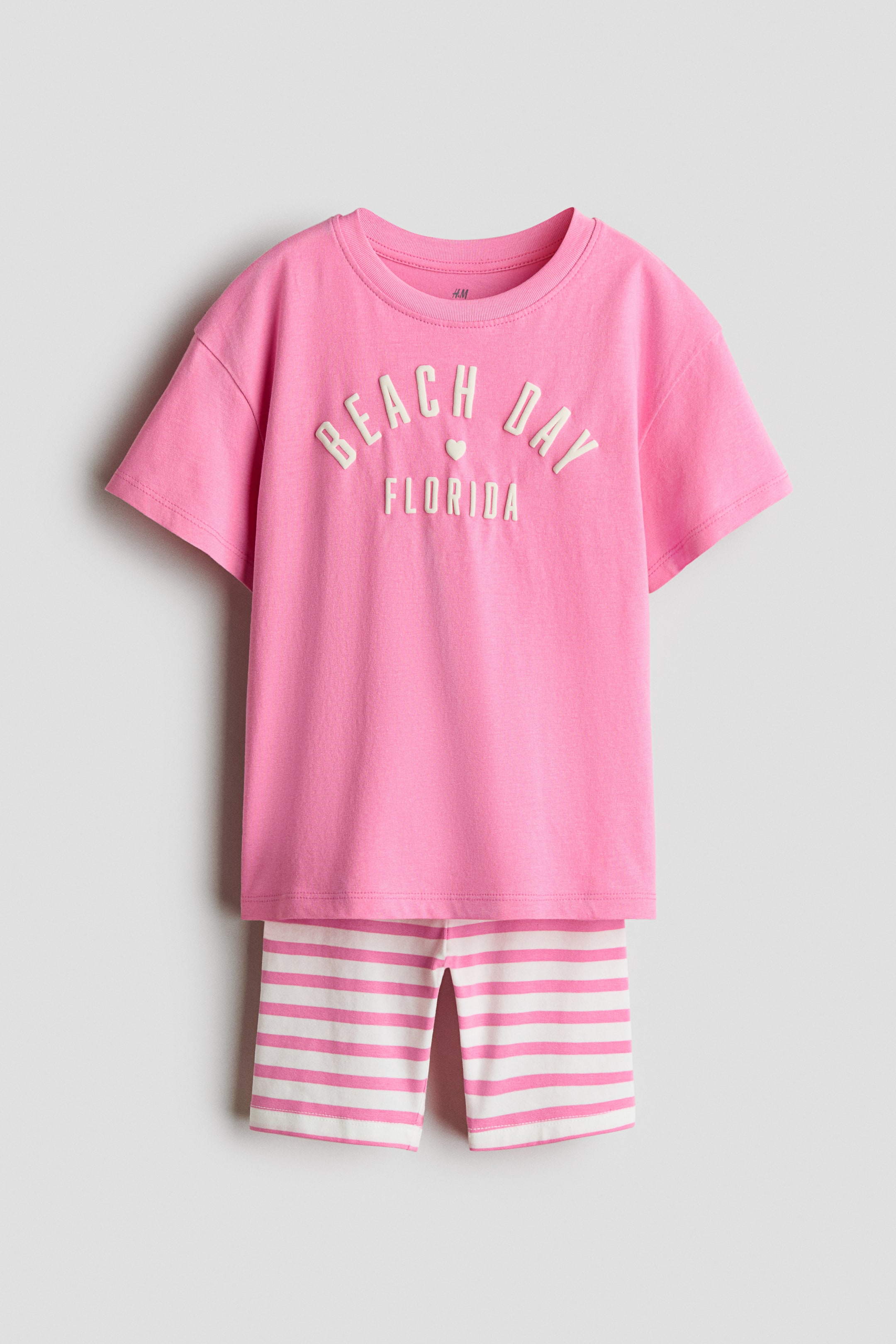 View larger image: A pink t-shirt and striped cycling shorts set is shown from the front. The t-shirt boasts a ribbed neckline, dropped shoulders, and raised "BEACH DAY FLORIDA" text with a heart, while the cycling shorts display white and pink horizontal stripes.