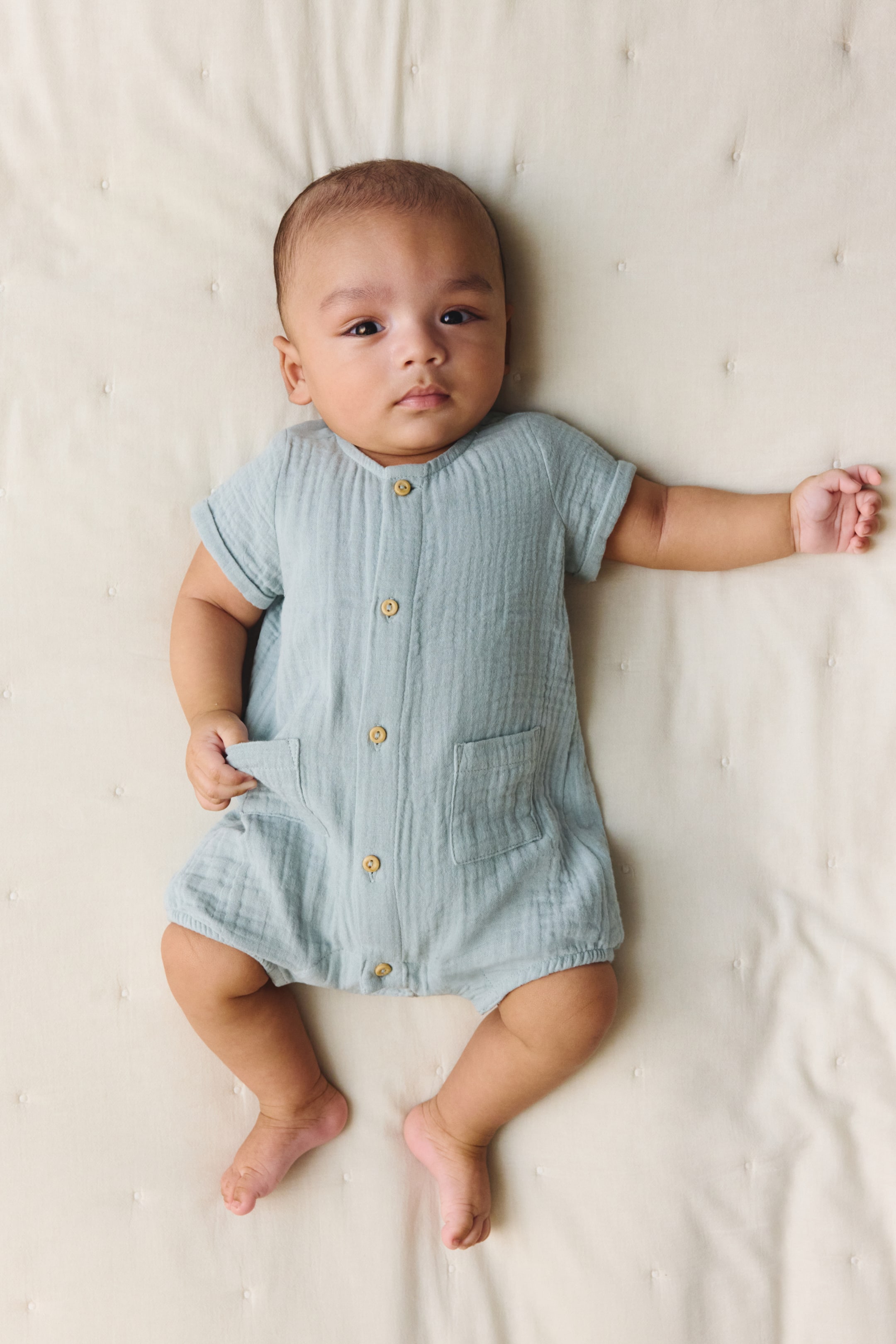 이미지 확대 보기: 연한 파란색 아기 롬퍼는 주름진 무슬린 원단으로 만들어졌으며, 둥근 목선과 연한 갈색 버튼이 있는 풀 버튼 다운 프론트를 특징으로 합니다. 짧고 롤업된 소매, 두 개의 앞 패치 포켓, 그리고 신축성 있는 다리 개구부로 디자인되었습니다.