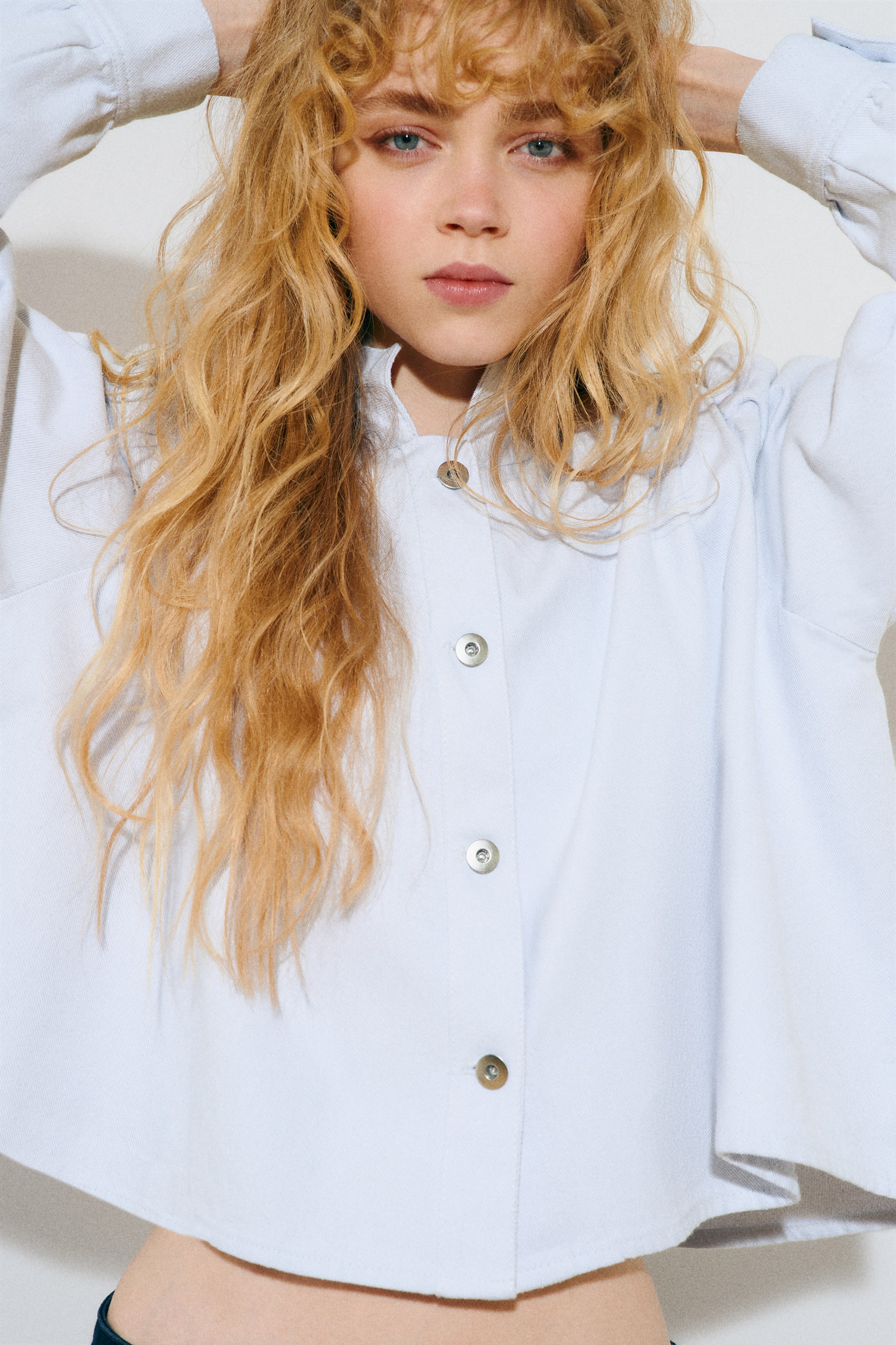 Προβολή μεγαλύτερης εικόνας: Η μοντέλο φοράει ένα ανοιχτό γαλάζιο κοντό πουκάμισο με όρθιο γιακά, κουμπιά σούστες κατά μήκος του μπροστινού μέρους και ένα V-σχήματος καρέ με πιέτες. Το πουκάμισο διαθέτει μακριά φουσκωτά μανίκια και η μοντέλο σηκώνει τα χέρια της, τονίζοντας τον όγκο των μανικιών.