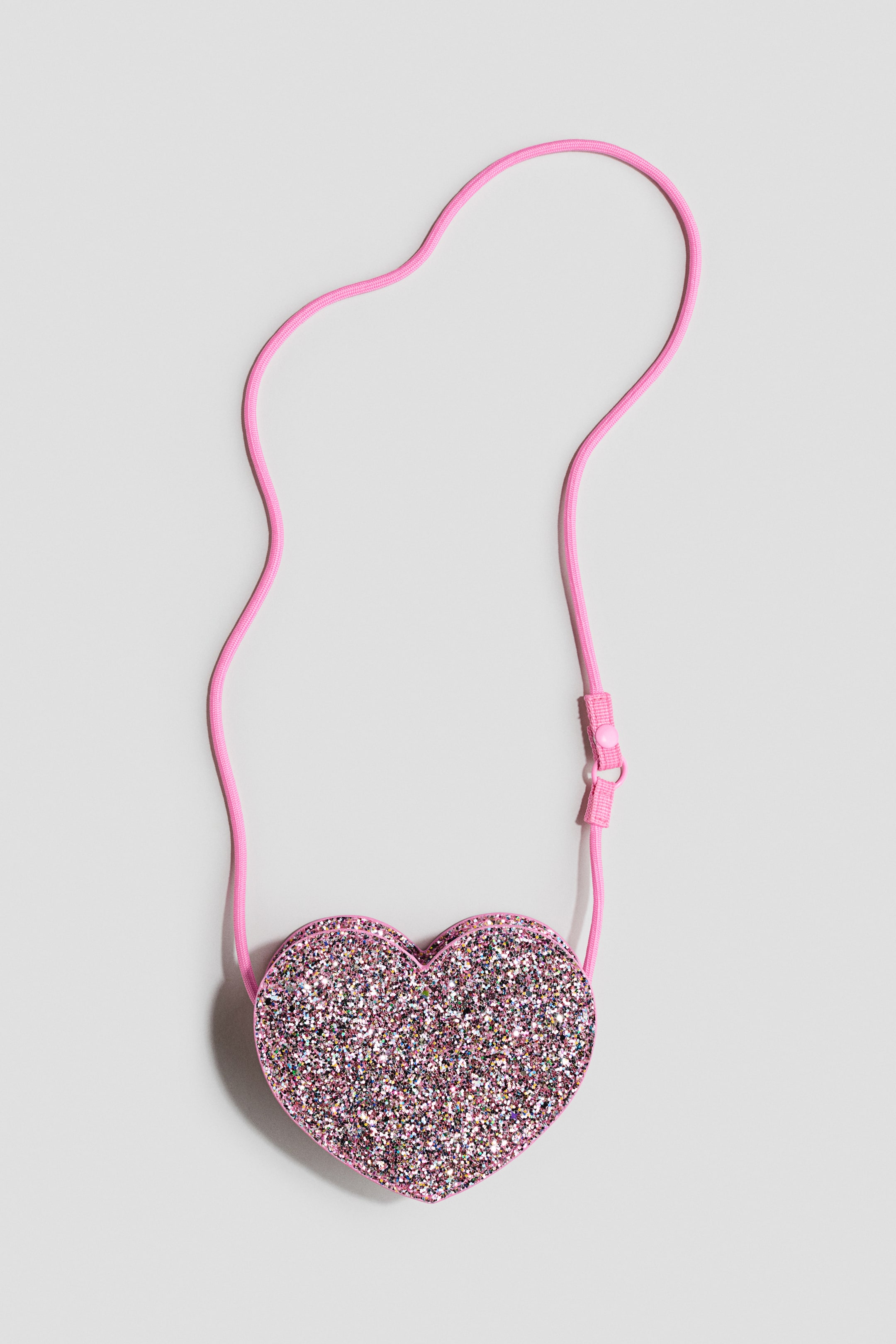 Voir une image plus grande: Un sac en forme de cœur présente une finition pailletée rose sur le devant, avec une fine sangle en cordon rose attachée aux côtés supérieurs et comprenant un petit ajusteur en plastique rose.