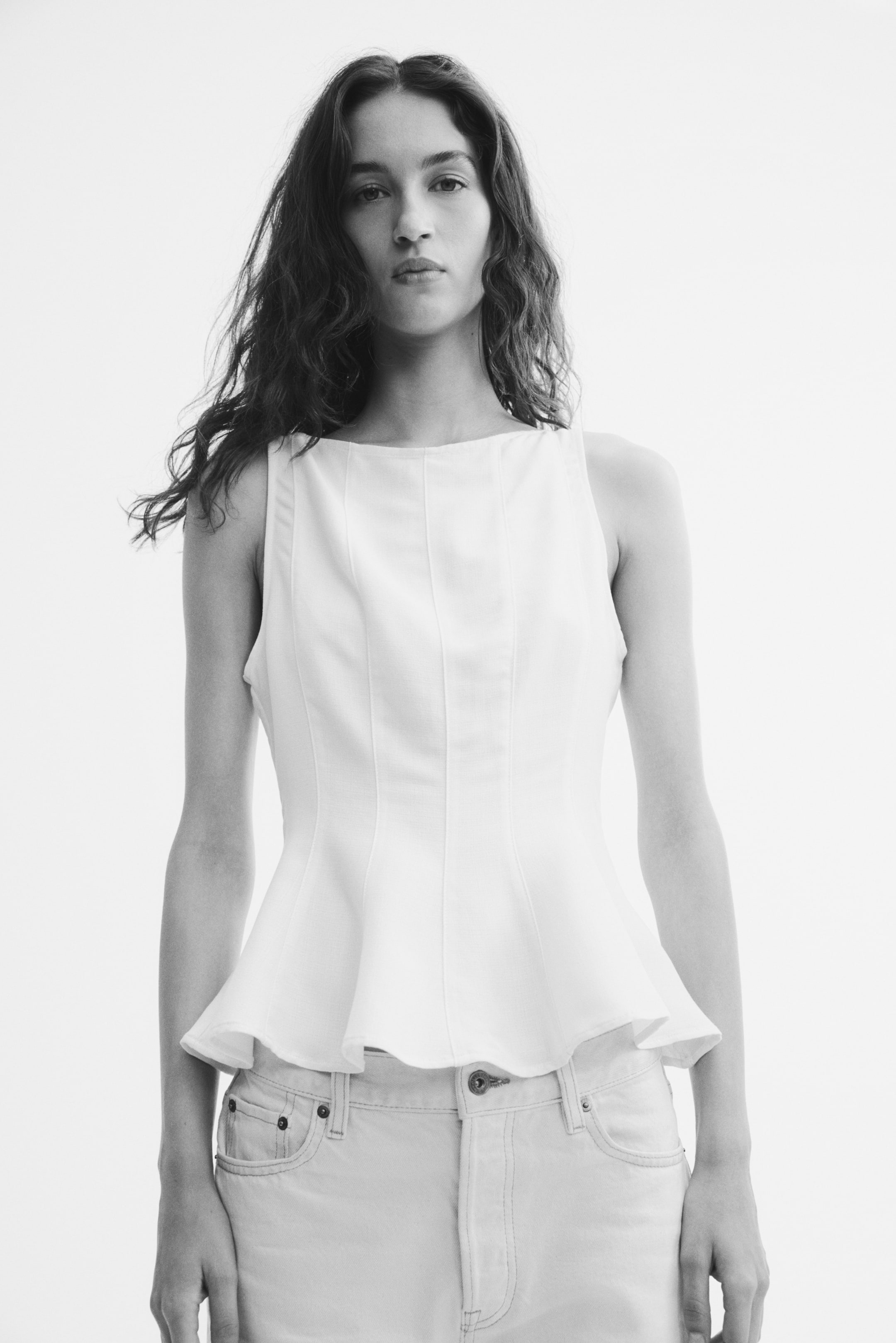 View larger image: The model is wearing a sleeveless, light-coloured peplum top with a boat neck and prominent vertical seams running down the front, styled with light-coloured jeans.