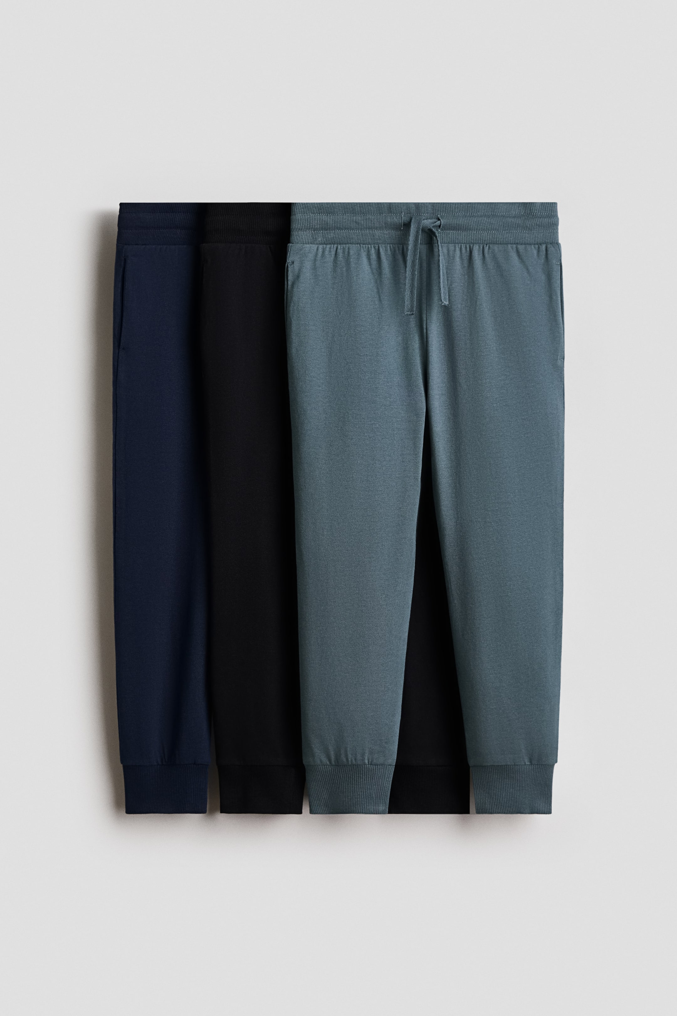 View larger image: Three pairs of cotton jersey joggers in navy blue, black, and muted blue-grey are hanging in a layered row, with the frontmost pair showing a front view. The joggers feature an elasticated, drawstring waistband, discreet side pockets, and ribbed cuffs.