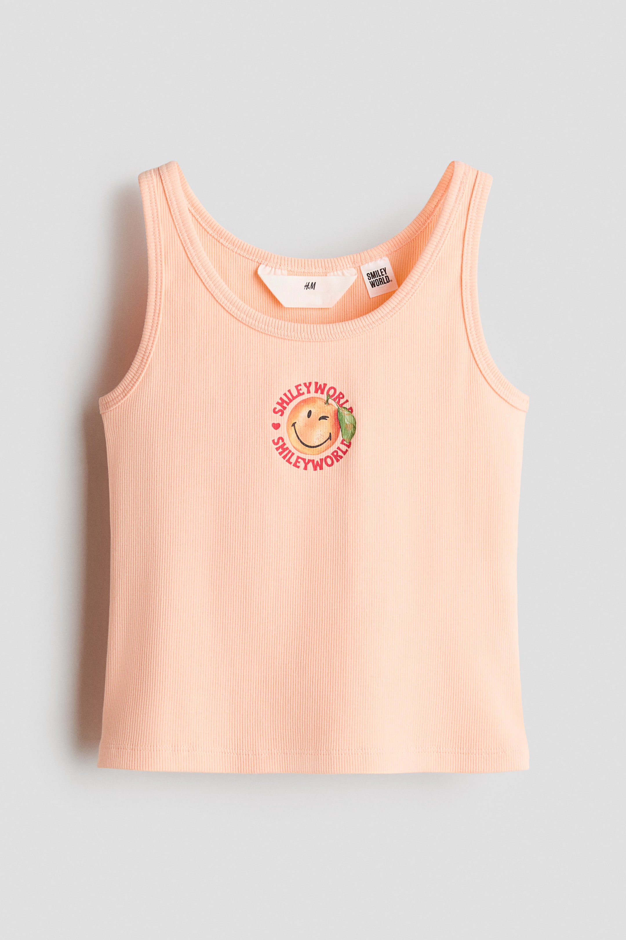 View larger image: A peach ribbed cotton jersey tank top is displayed from the front, showcasing a circular print motif on the chest featuring a winking smiley face inside a peach fruit, surrounded by the text "SMILEY WORLD." Narrow trims detail the round neckline and armholes.
