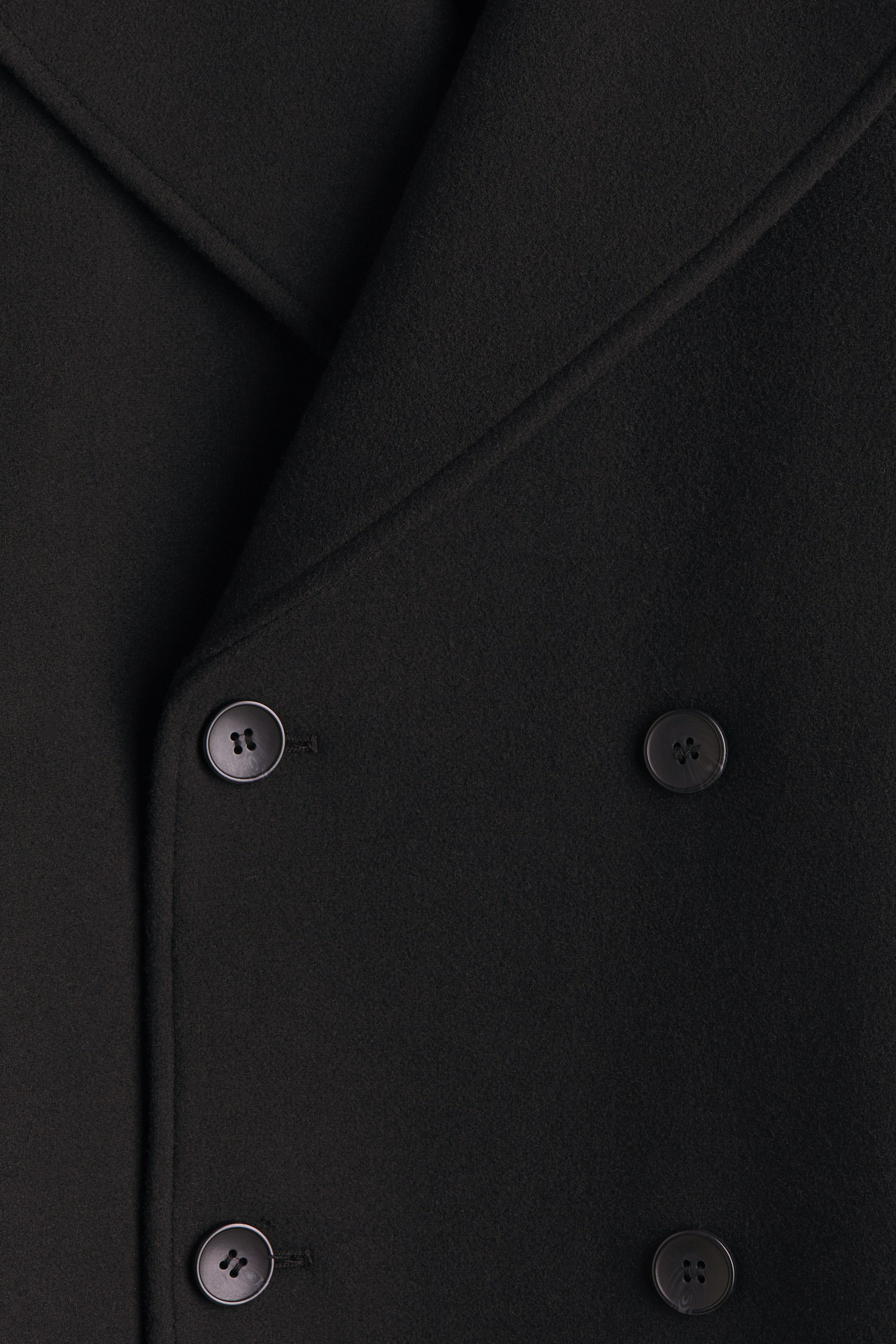 拡大画像を表示: 黒のダブルブレストコートの前面には、4つの暗い色の丸い四つ穴ボタンが2列に並び、細かい質感の生地の上に幅広のラペルが折り重なっています。