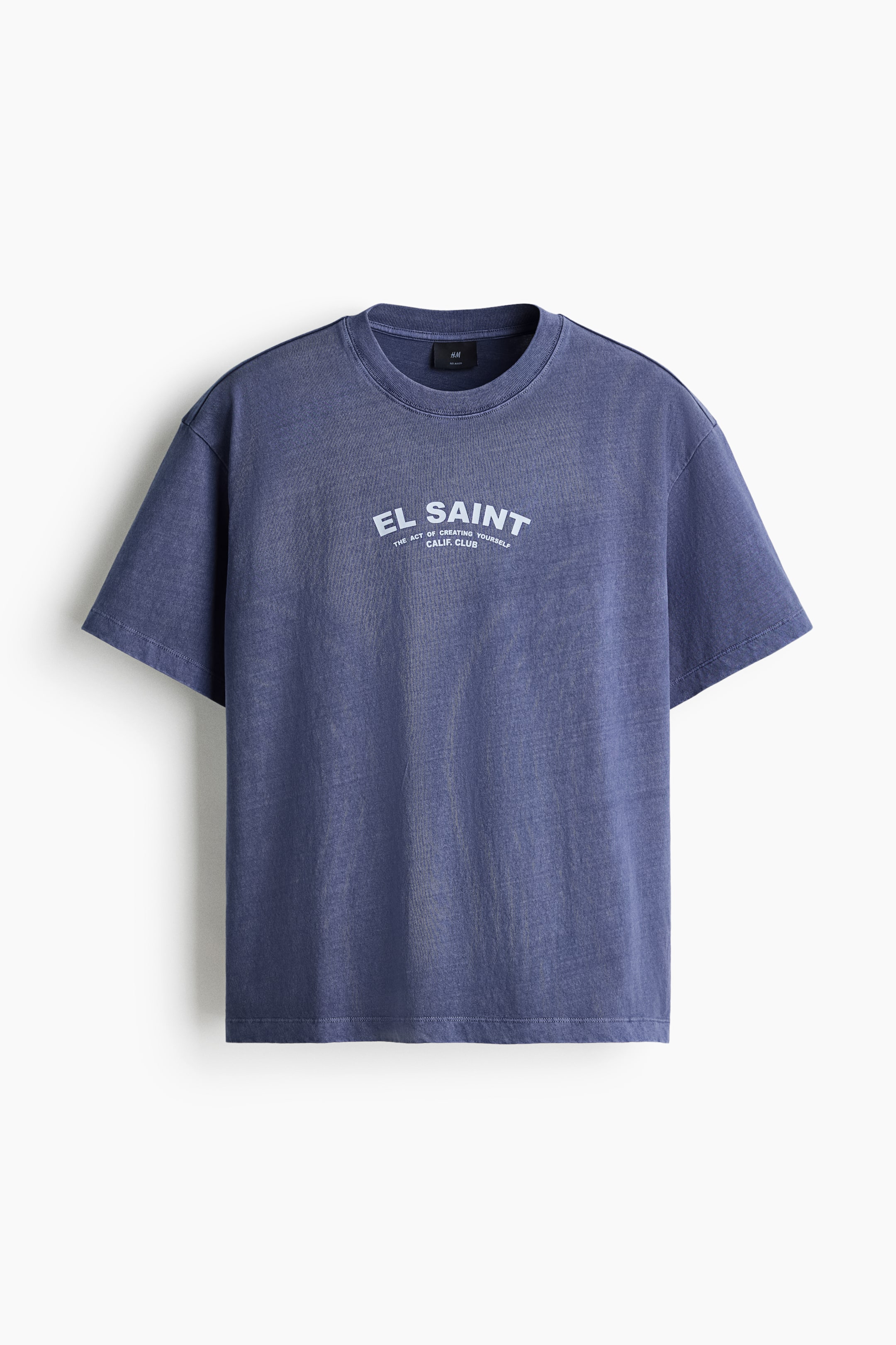 View larger image: A washed blue short-sleeved t-shirt is displaying its front, featuring a ribbed crew neck and a white graphic print on the chest that reads 'EL SAINT' with smaller text below.