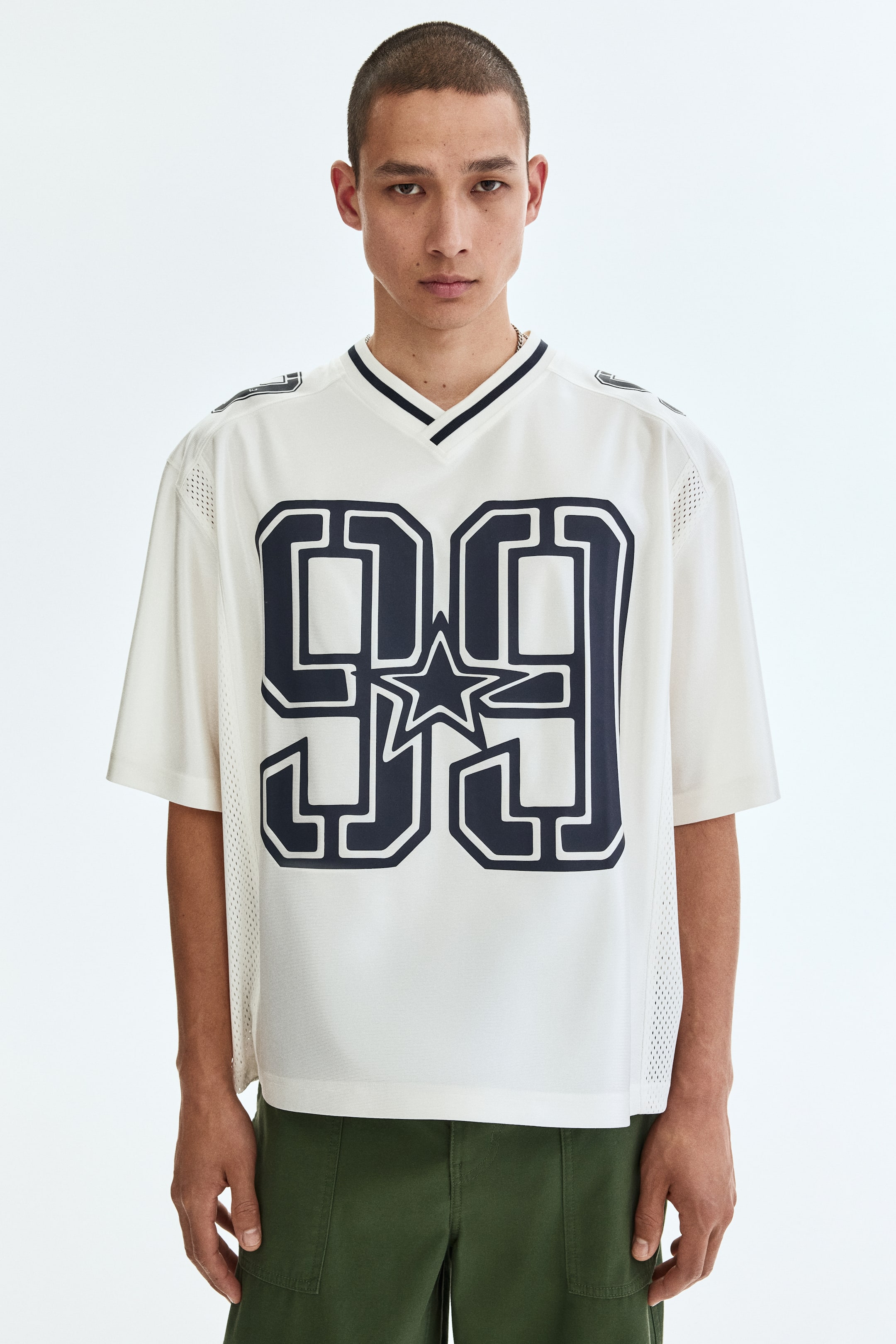 Agrandir l'image: Un T-shirt style boxy en jersey mesh de couleur blanc cassé, arborant un grand imprimé '99' bleu foncé avec une étoile et un col en V côtelé, est porté par le modèle. Le T-shirt présente des épaules tombantes et un ourlet droit, offrant une coupe ample.