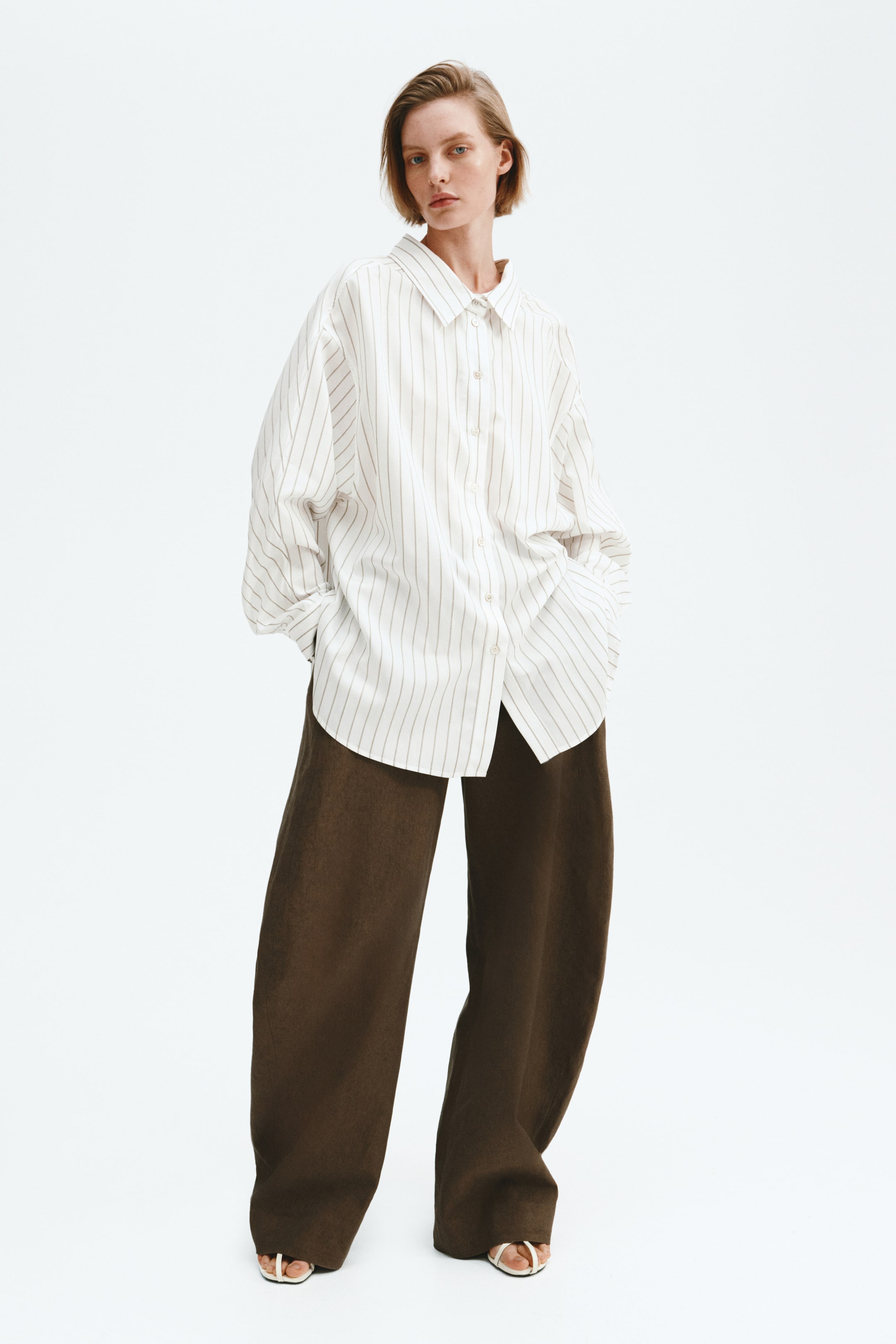 View larger image: A white oversized button-down shirt with thin vertical stripes is showcased from the front, featuring a pointed collar and long sleeves with rolled-up cuffs. Dark brown wide-leg trousers are worn underneath, cascading over white open-toe sandals.