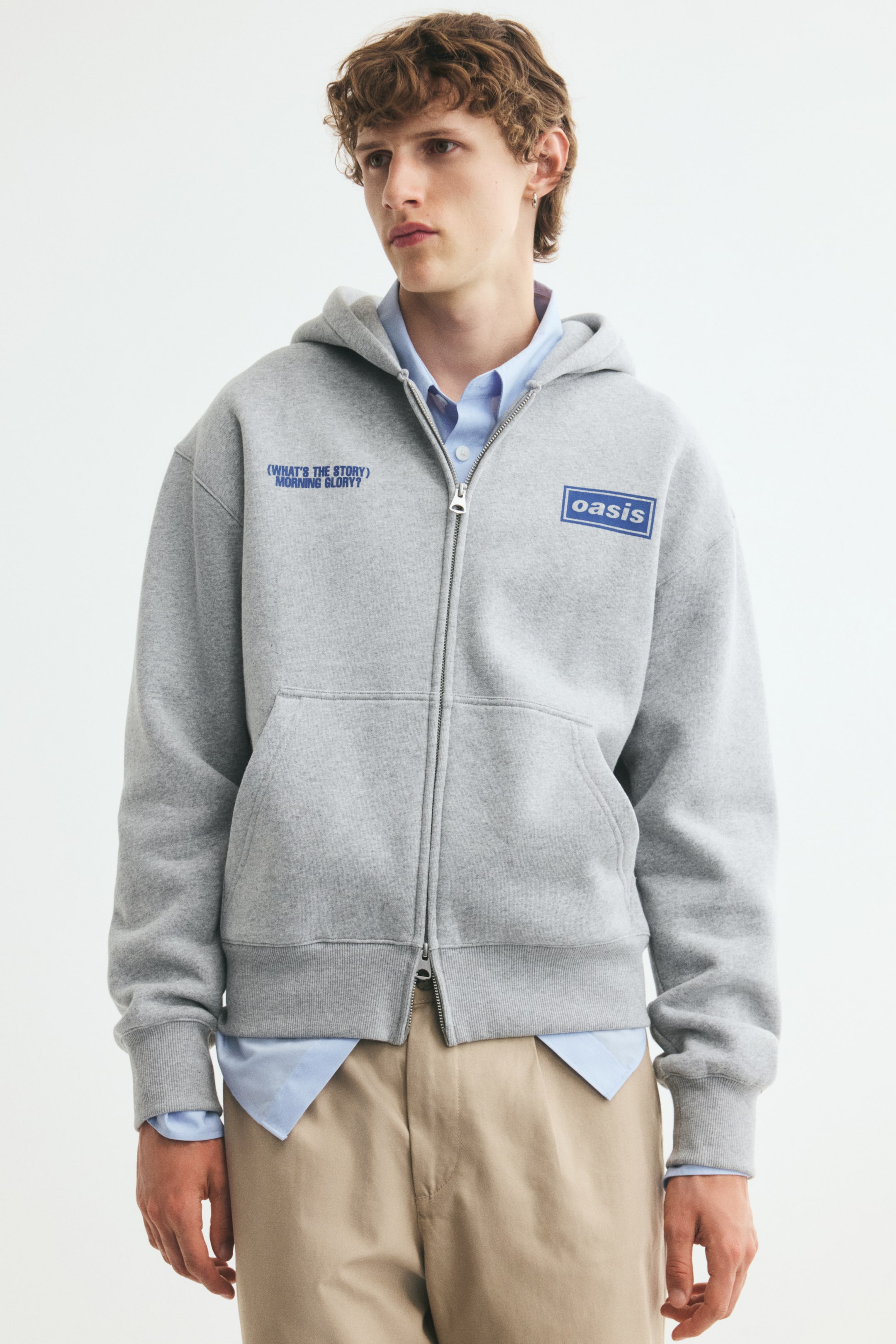 View larger image: The model is wearing a grey zip-up hoodie over a light blue collared shirt and beige trousers. The hoodie features blue printed graphics on the chest, a partially zipped two-way front zipper, split front pockets, dropped shoulders, and ribbed cuffs and hem.