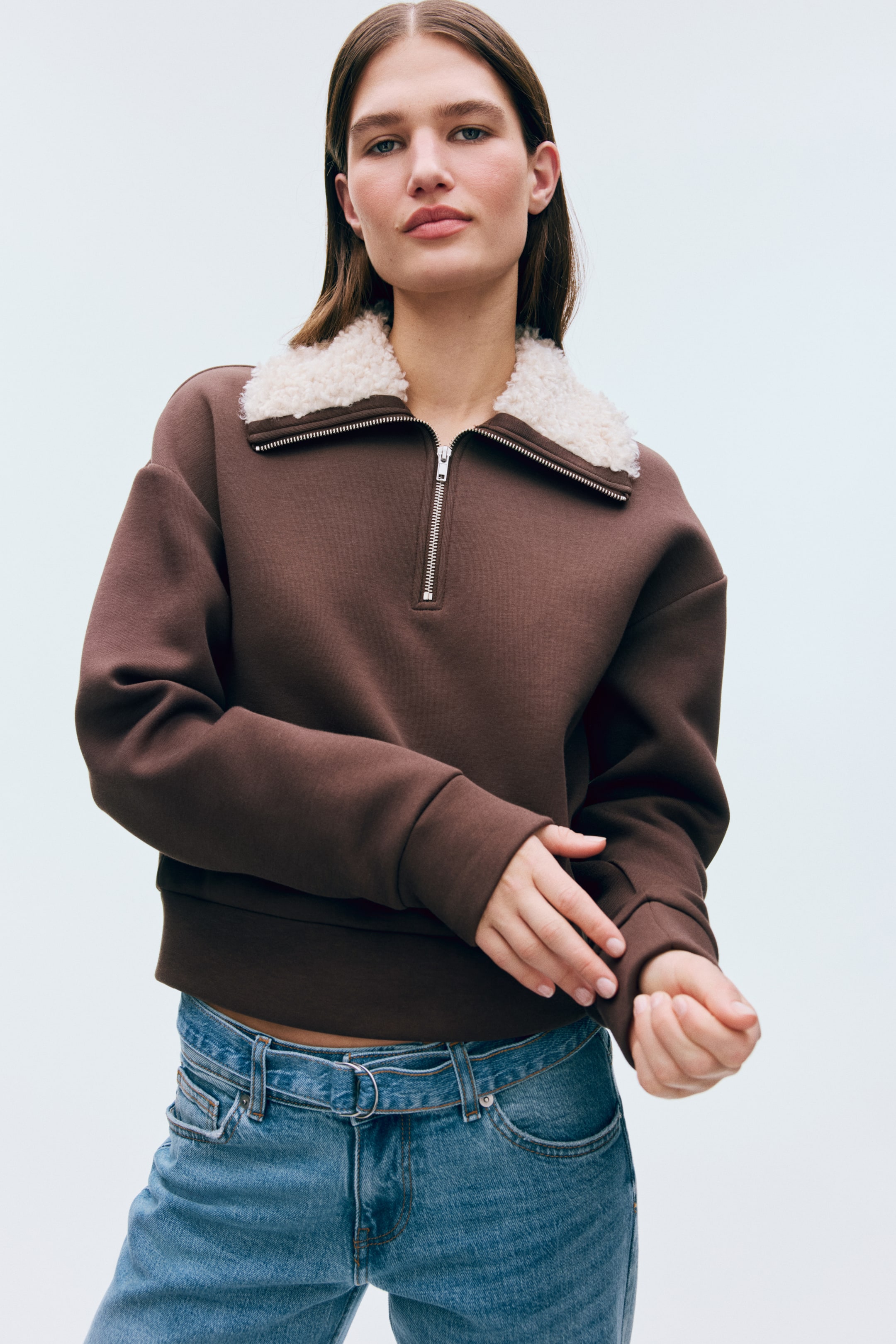 View larger image: A brown loose-fit sweatshirt in scuba fabric is worn by the model, who is adjusting one of its wide cuffs. It features a teddy-lined stand collar with a top zipper, dropped shoulders, and wide trims at the cuffs and hem.