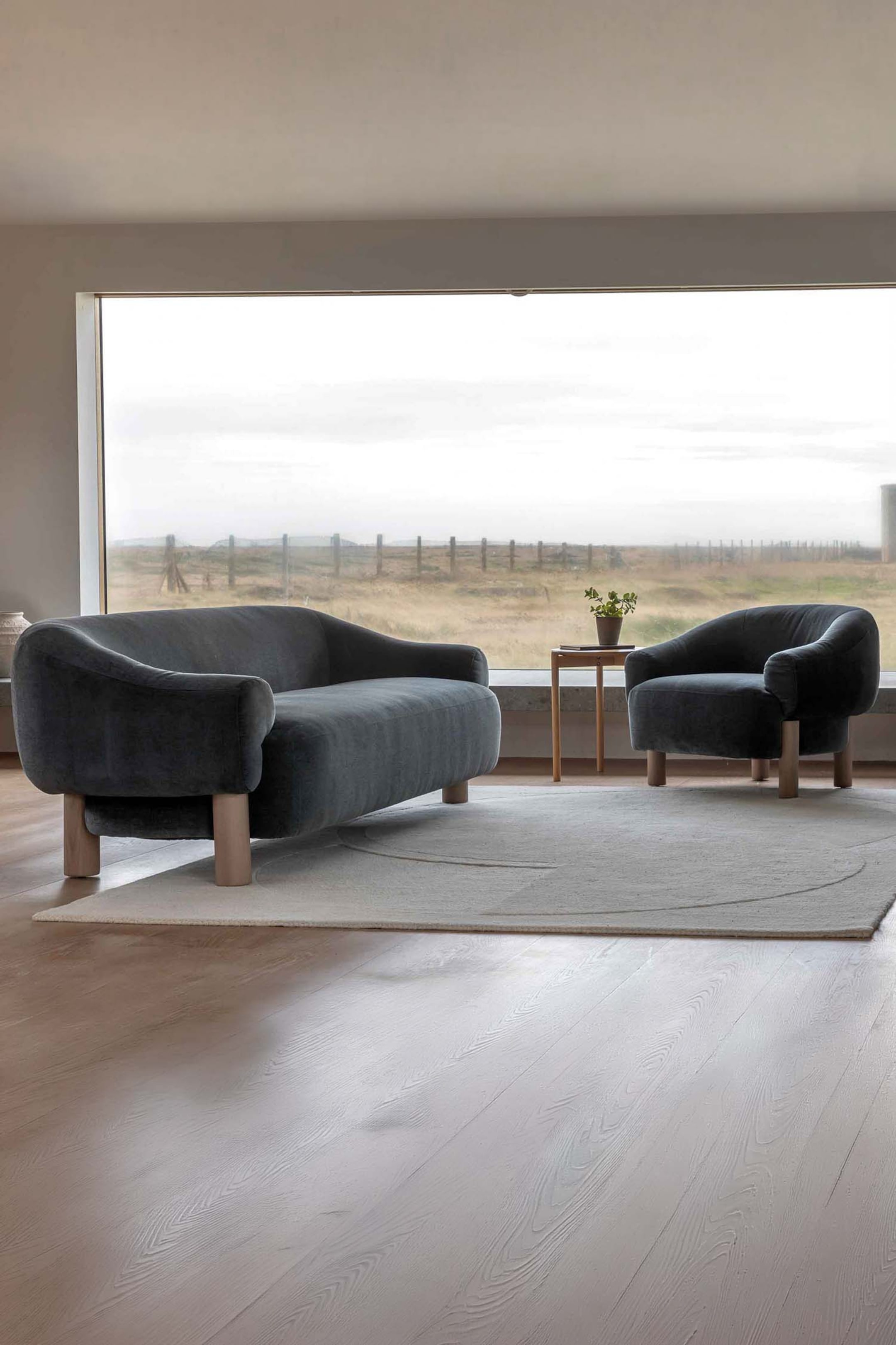 View larger image: A dark grey upholstered sofa and a matching armchair are showcasing rounded, low-profile designs. Both pieces feature thick, light wooden legs, with the sofa angled slightly to the right and the armchair angled slightly to the left, highlighting their curved forms.