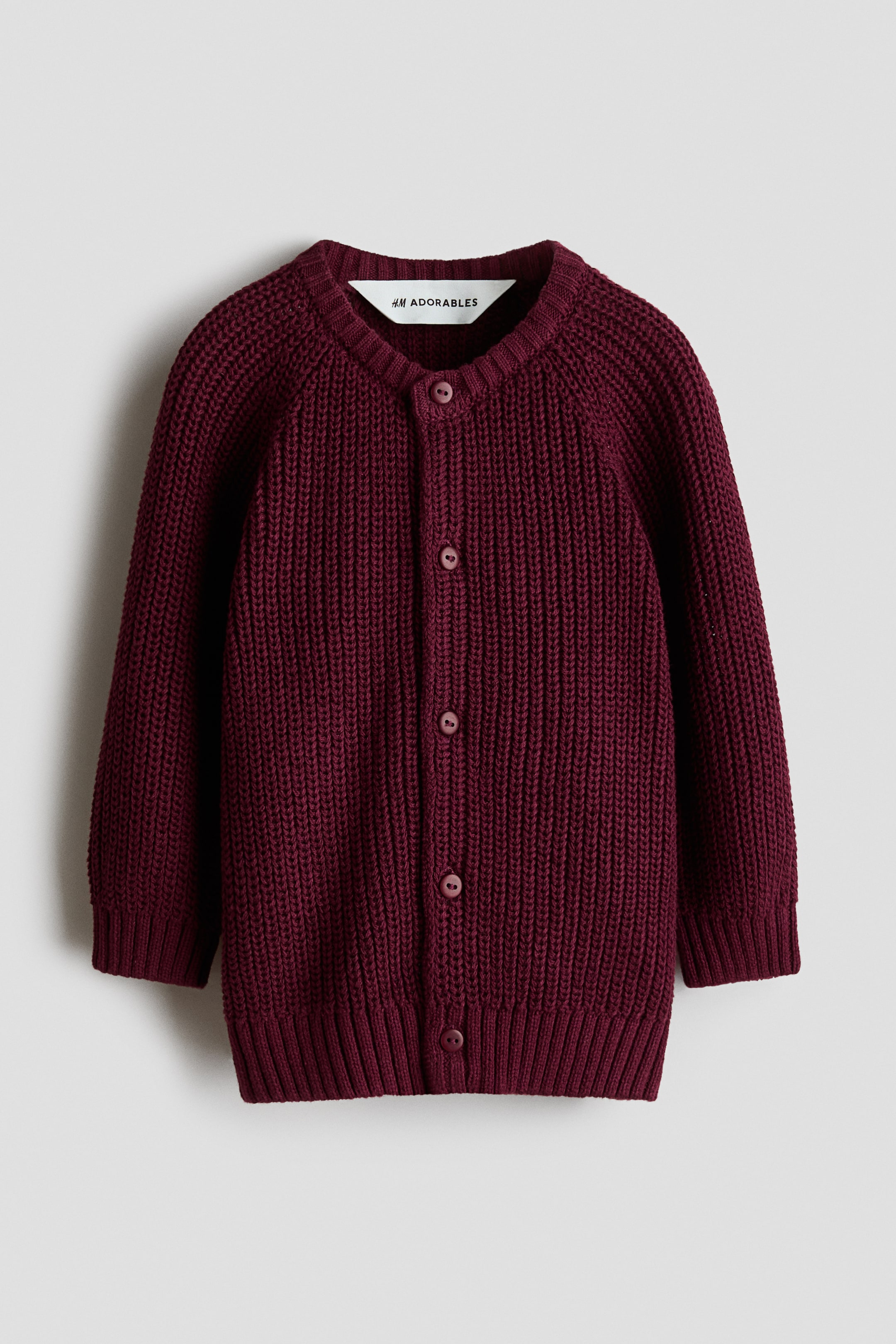 Se større billede: En bordeaux cardigan vises forfra og fremhæver en kraftig strikket tekstur, en rund halsudskæring med ribkant og lange raglanærmer. Ribkanter detaljerer manchetterne og kanten, og fem runde knapper lukker forstykket.