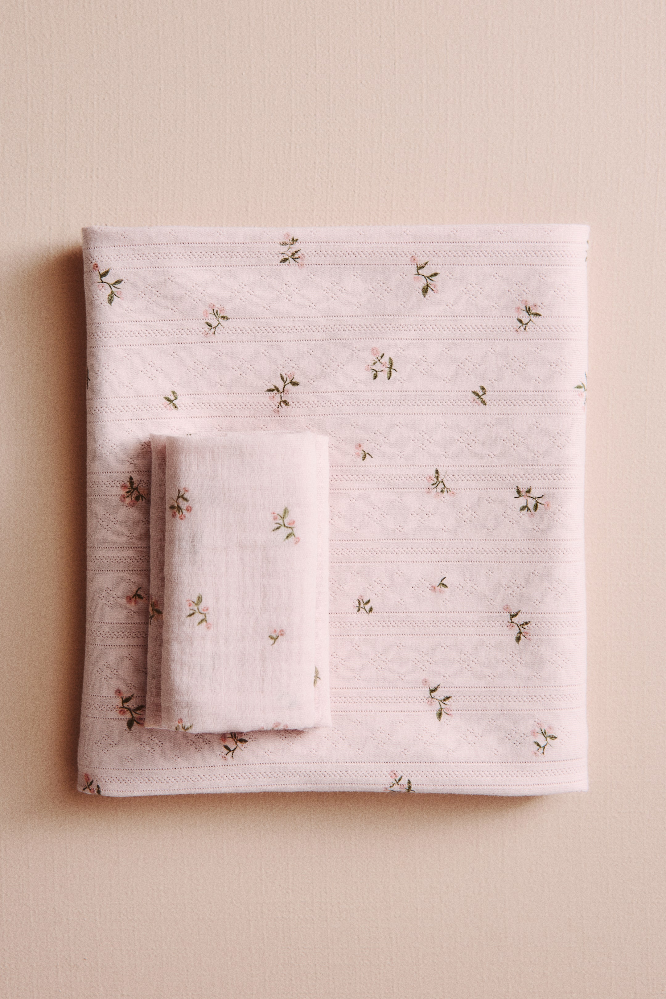 Voir une image plus grande: Une couverture pour bébé en coton tricoté pointelle rose arbore un imprimé floral classique de petites fleurs roses et de feuilles vertes. Un textile plus petit, plié, avec un imprimé floral assorti repose sur le dessus.