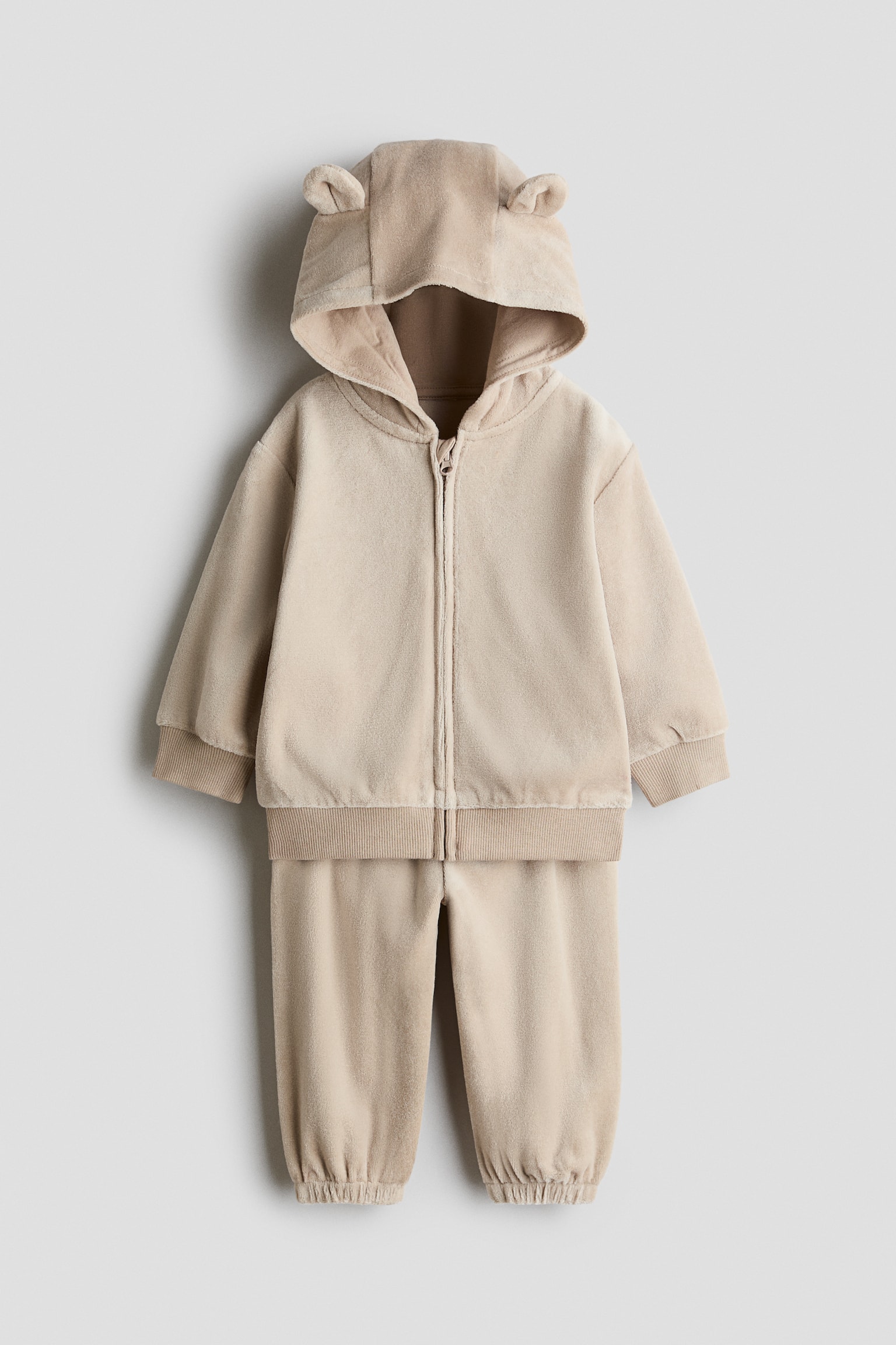 A beige velour two-piece baby outfit features a front-facing zip-up hoodie with small rounded ears on the hood, ribbed cuffs, and a ribbed hem. The matching trousers complete the set with a relaxed fit and elasticated ankle cuffs.