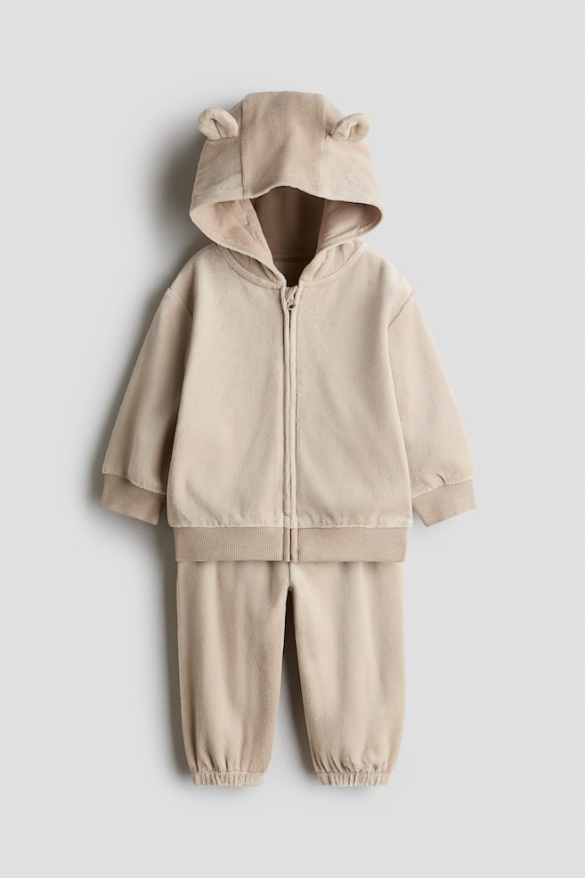 Un completo per neonato in velluto beige composto da due pezzi, con una felpa con cappuccio e zip frontale dotata di piccole orecchie arrotondate sul cappuccio, polsini a costine e orlo a costine. I pantaloni coordinati completano il set con una vestibilità comoda e fondo caviglia elasticizzato. - 1