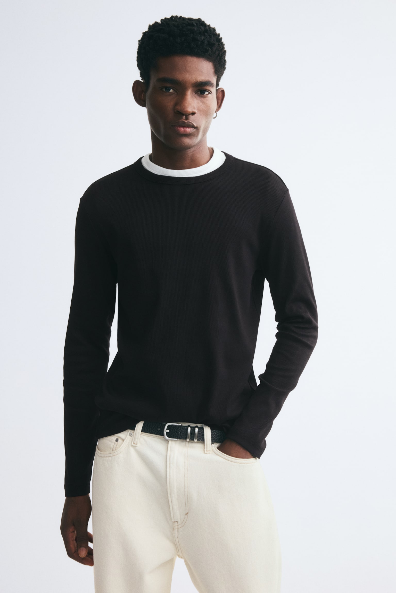 A dark brown long-sleeve tee in slim-fit ribbed cotton jersey is worn by the model, layered over a white T-shirt visible at the round neckline. The tee is styled with cream-coloured jeans and a black belt. - 1