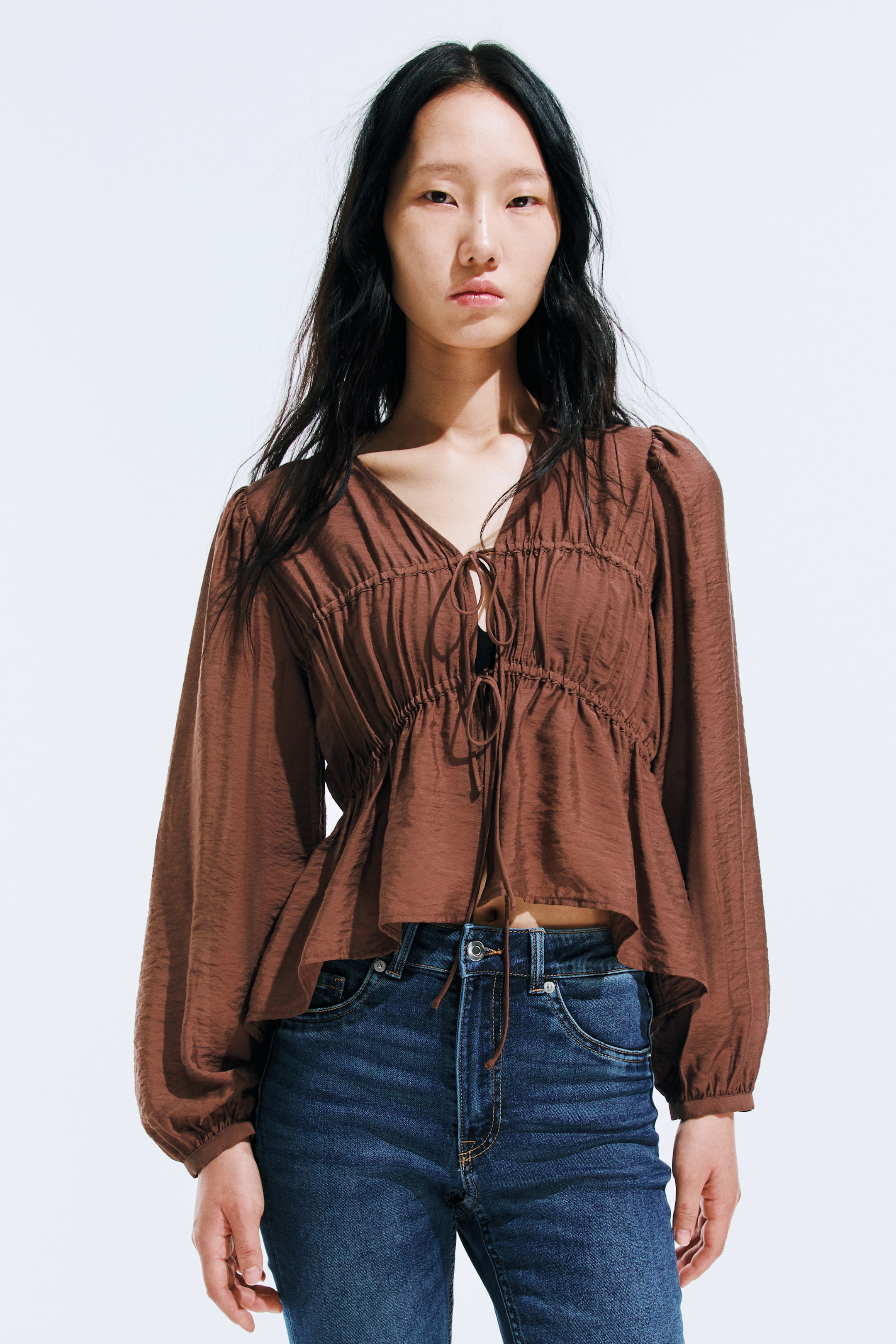 Xem ảnh lớn hơn: Một chiếc áo blouse nâu dáng lửng nổi bật với cổ chữ V và chi tiết buộc dây phía trước, cùng với những đường xếp ly dày đặc trên thân áo, tạo hiệu ứng kết cấu và ôm sát. Thiết kế với tay áo dài, phồng, kết thúc bằng cổ tay áo xếp ly và đường viền xòe.