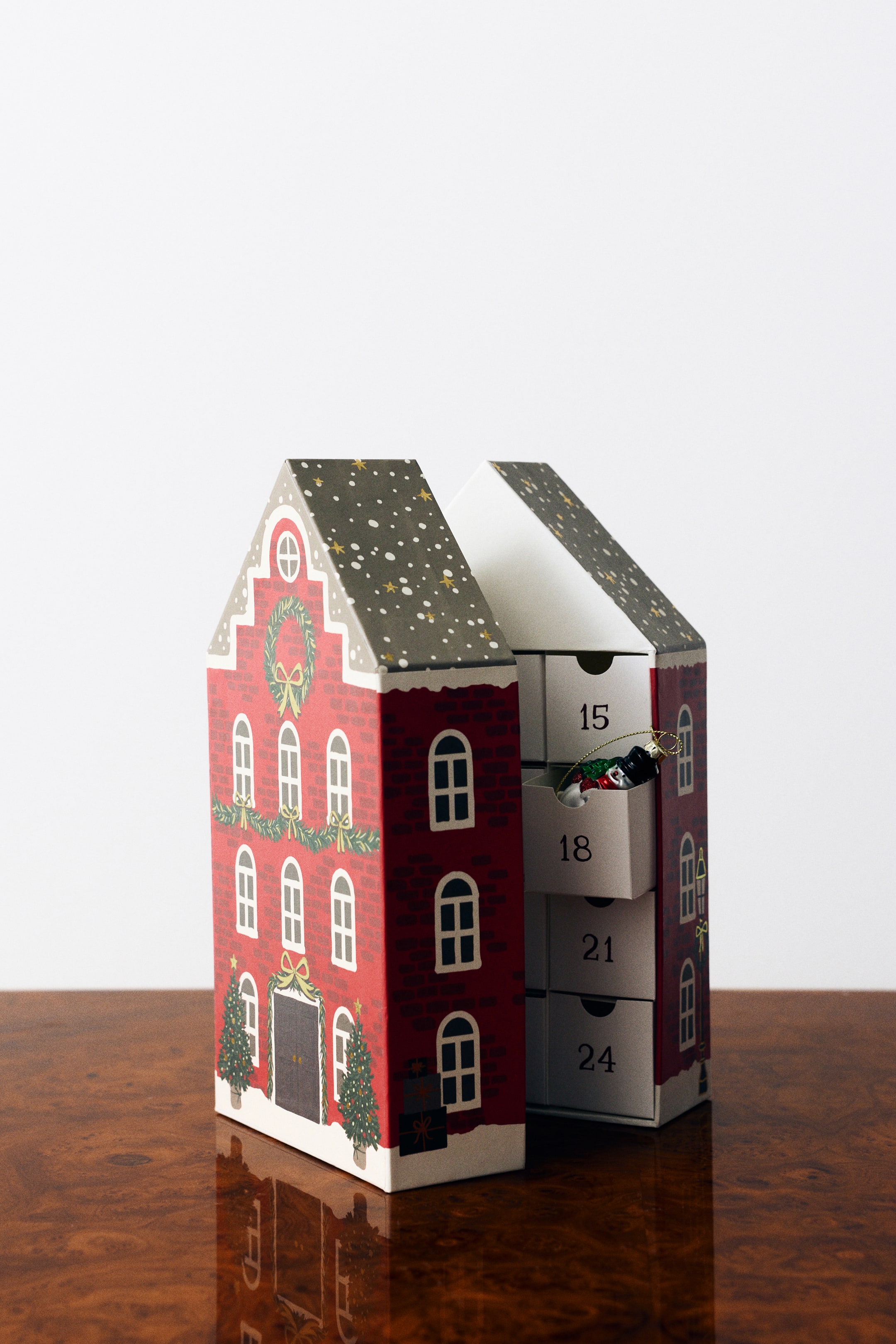 View larger image: A house-shaped Advent calendar is shown slightly open from a three-quarter angle, featuring a red-brick facade decorated with green wreaths and Christmas trees on the left side. The right side reveals a column of white numbered drawers, with drawer number 18 partially pulled open to display a small festive ornament inside.