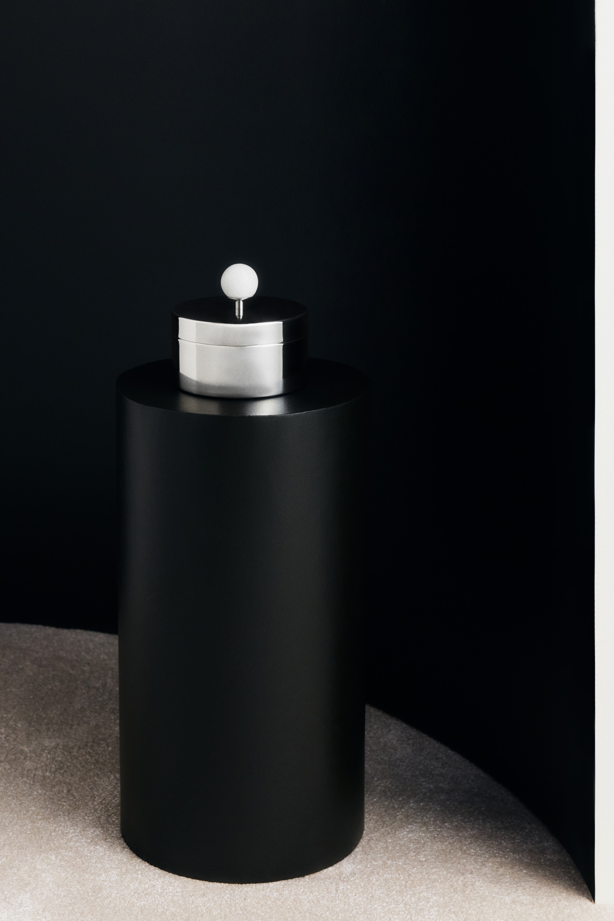 A polished silver-toned metal container is shown from the front. It is featuring a cylindrical body and a flat lid, with a white spherical marble knob rising centrally from the lid on a slender metal stem.
