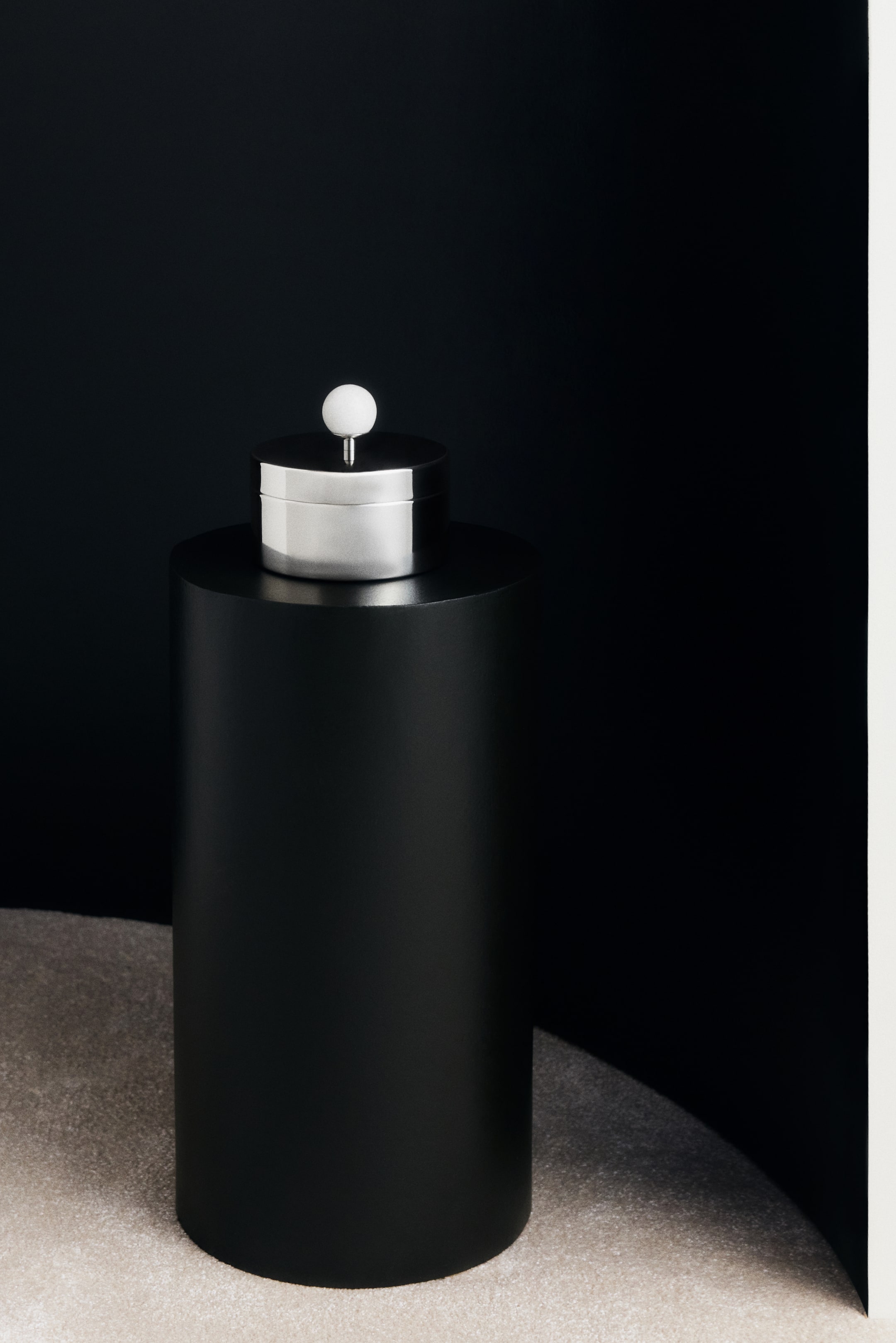 Voir une image plus grande: Une boîte ronde en acier inoxydable repose sur un piédestal cylindrique noir, vue de face. La boîte est dotée d’un couvercle plat assorti, surmonté d’un bouton sphérique en marbre blanc fixé sur une courte tige métallique.