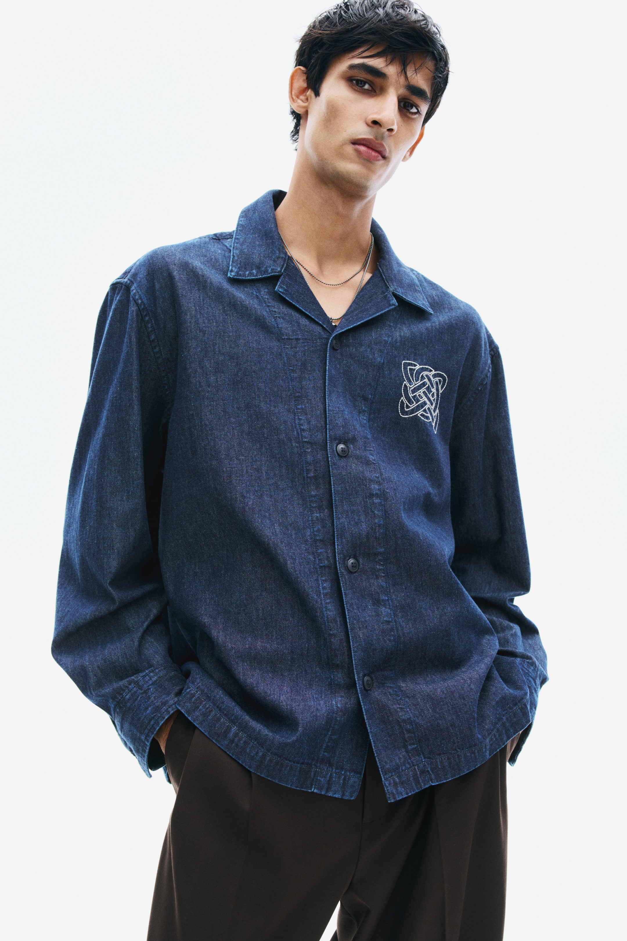 View larger image: A dark blue denim overshirt featuring a resort collar, button closures, and a white embroidered knot detail on the chest is being worn by the model. The model is posing with hands in the pockets of dark brown trousers, showcasing the shirt's straight hem and long sleeves.