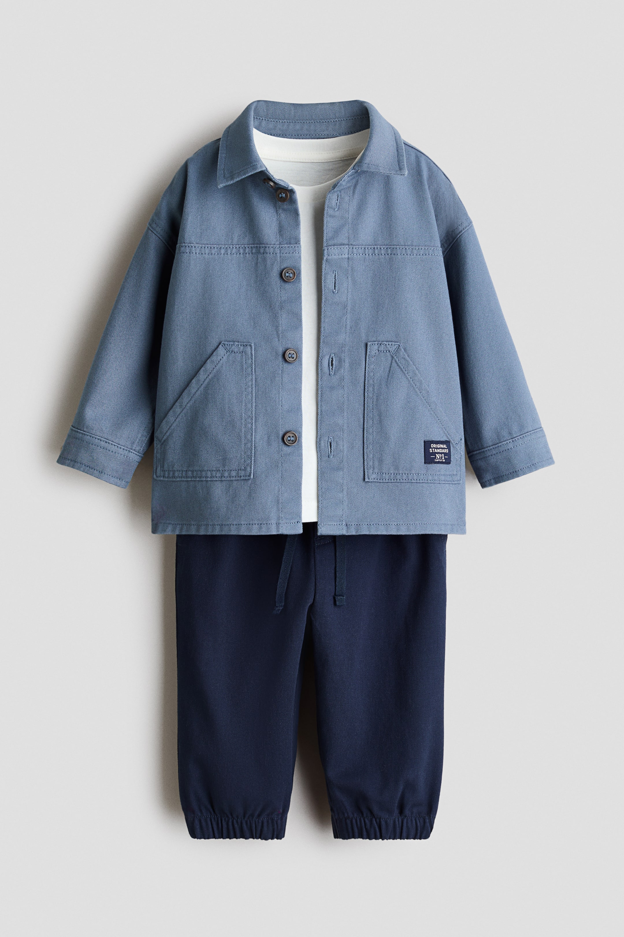 View larger image: A blue overshirt featuring a pointed collar, a brown button-front closure, and two large front patch pockets, with one pocket displaying a small rectangular label. This overshirt is layered over a white t-shirt, and dark blue jogger pants are visible below, showcasing an elasticated drawstring waist and elasticated cuffs. The full set is facing front.