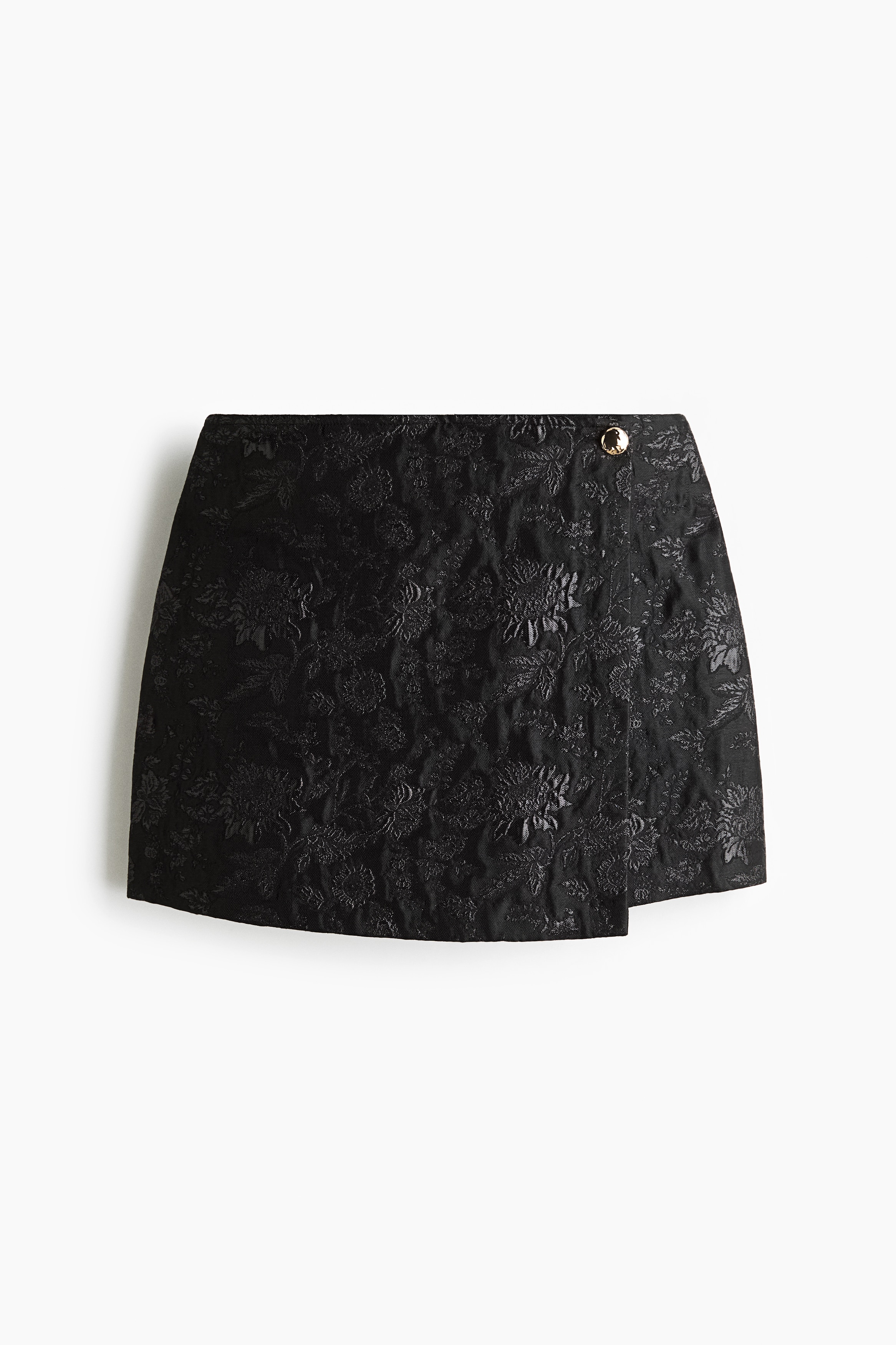 A black jacquard wrap skirt is shown from the front, displaying a textured floral pattern and an asymmetrical wrap design. The skirt features a flat waistband secured with a single decorative gold-tone button on the upper right side.
