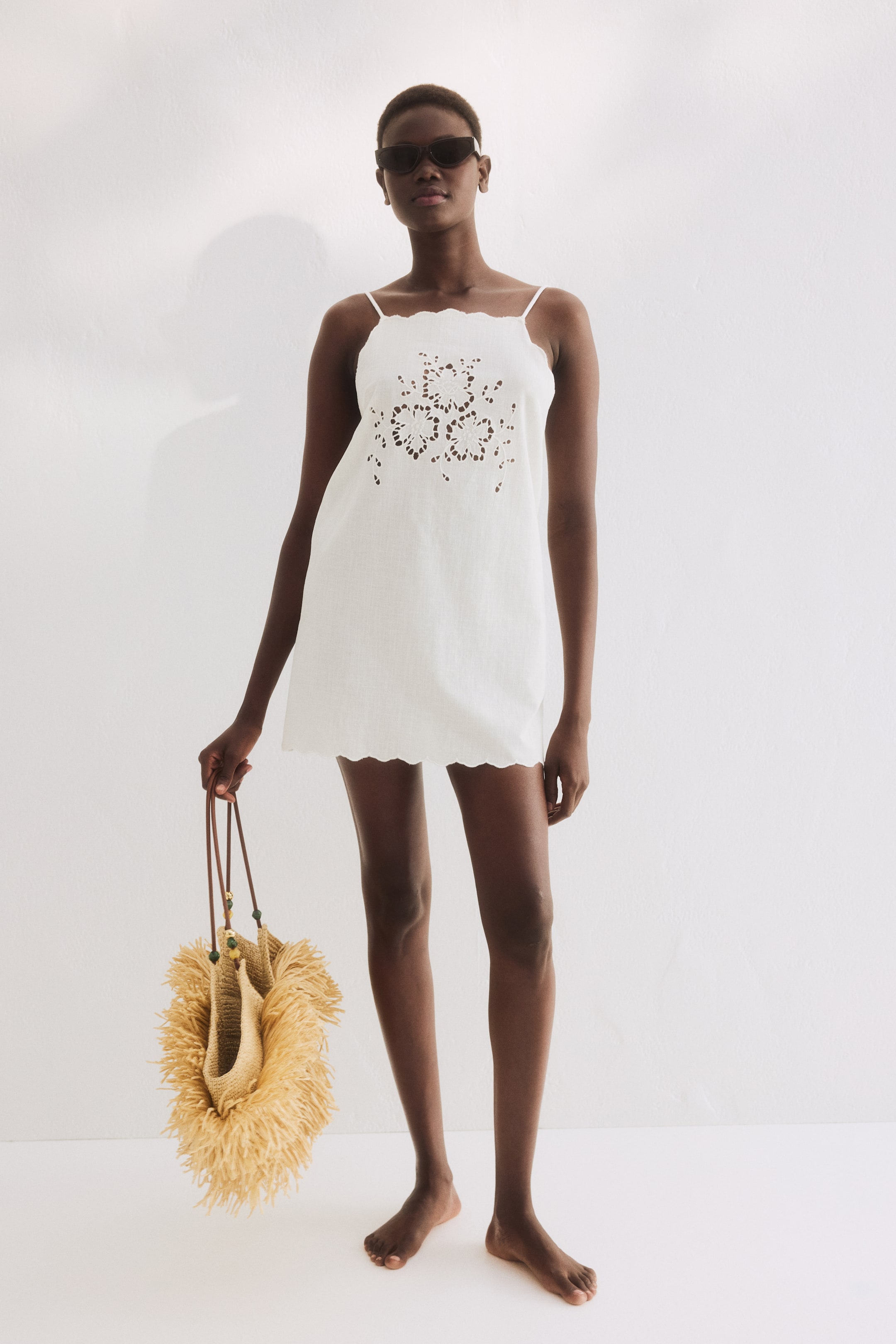 View larger image: The model is wearing a short beach dress crafted from an airy cotton weave, highlighted by a straight neckline, narrow shoulder straps, broderie anglaise detailing at the front, and scalloped edges on the hem and neckline. She's also holding a fringed straw bag.