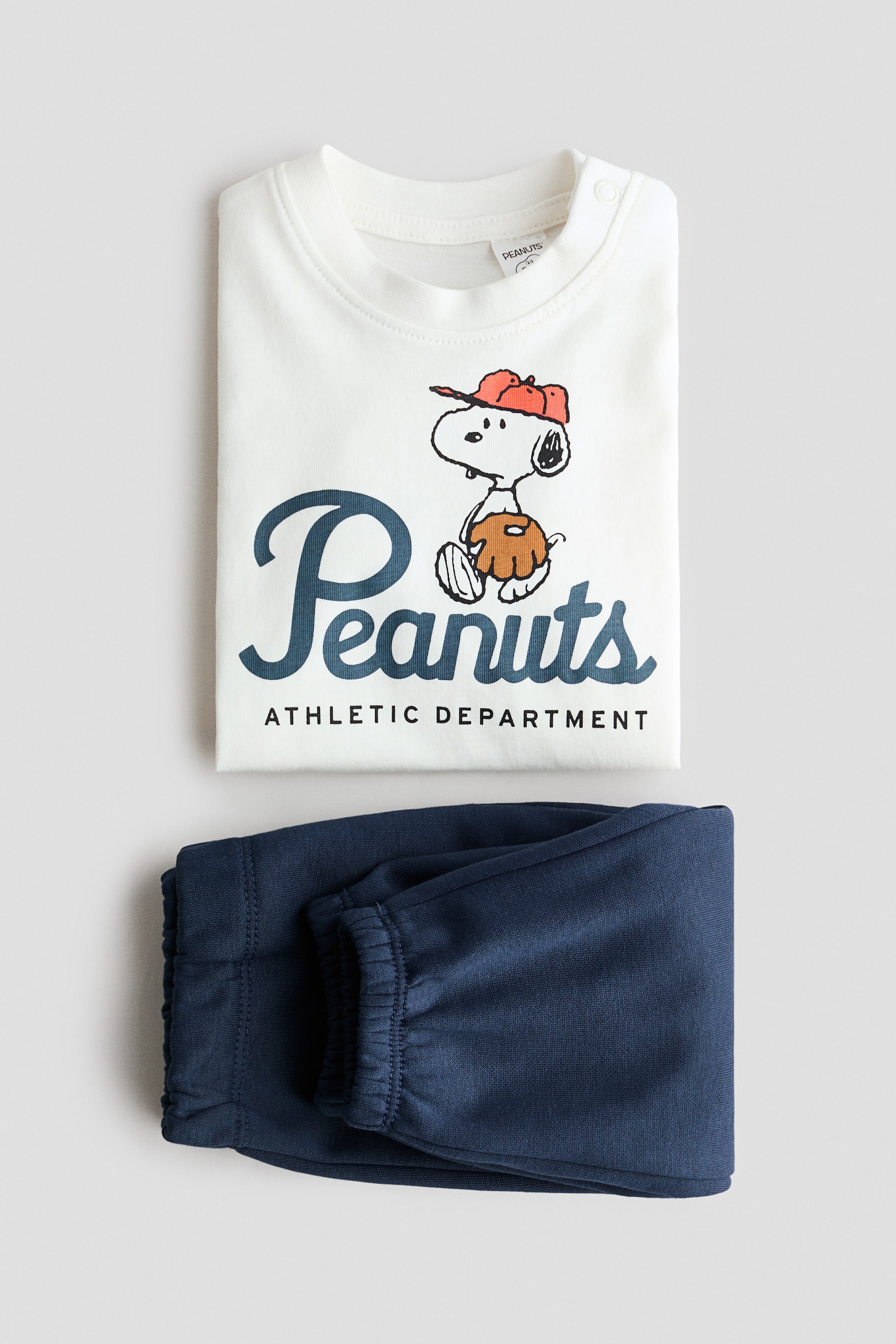 Visualizza immagine più grande: Un set di abbigliamento per neonati composto da due pezzi è presentato, caratterizzato da un top a maniche lunghe color bianco sporco con scollo rotondo, bottoni a pressione sulle spalle e una grafica frontale di Snoopy con cappello rosso e guanto marrone, accompagnata dal testo 'Peanuts Athletic Department'. Un paio di pantaloni stile jogger di colore blu scuro con polsini elasticizzati è piegato ordinatamente sotto il top.