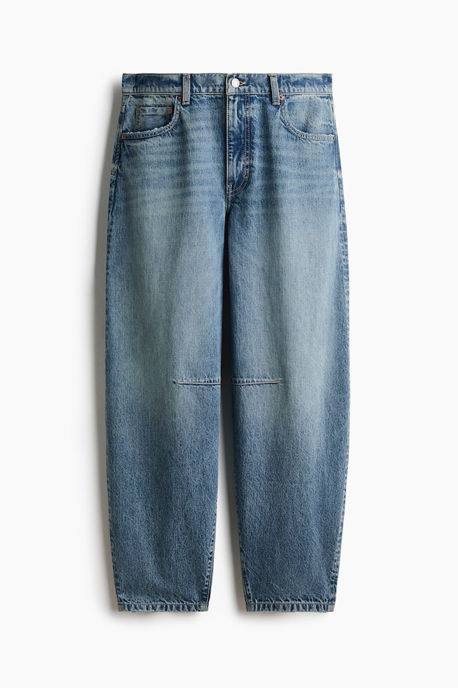 A pair of medium-wash blue denim jeans is hanging front-facing, featuring a voluminous, curved-leg silhouette that tapers towards the hem. The jeans feature horizontal knee darts, classic five-pocket styling, and a regular waist with a button fastening. - 1