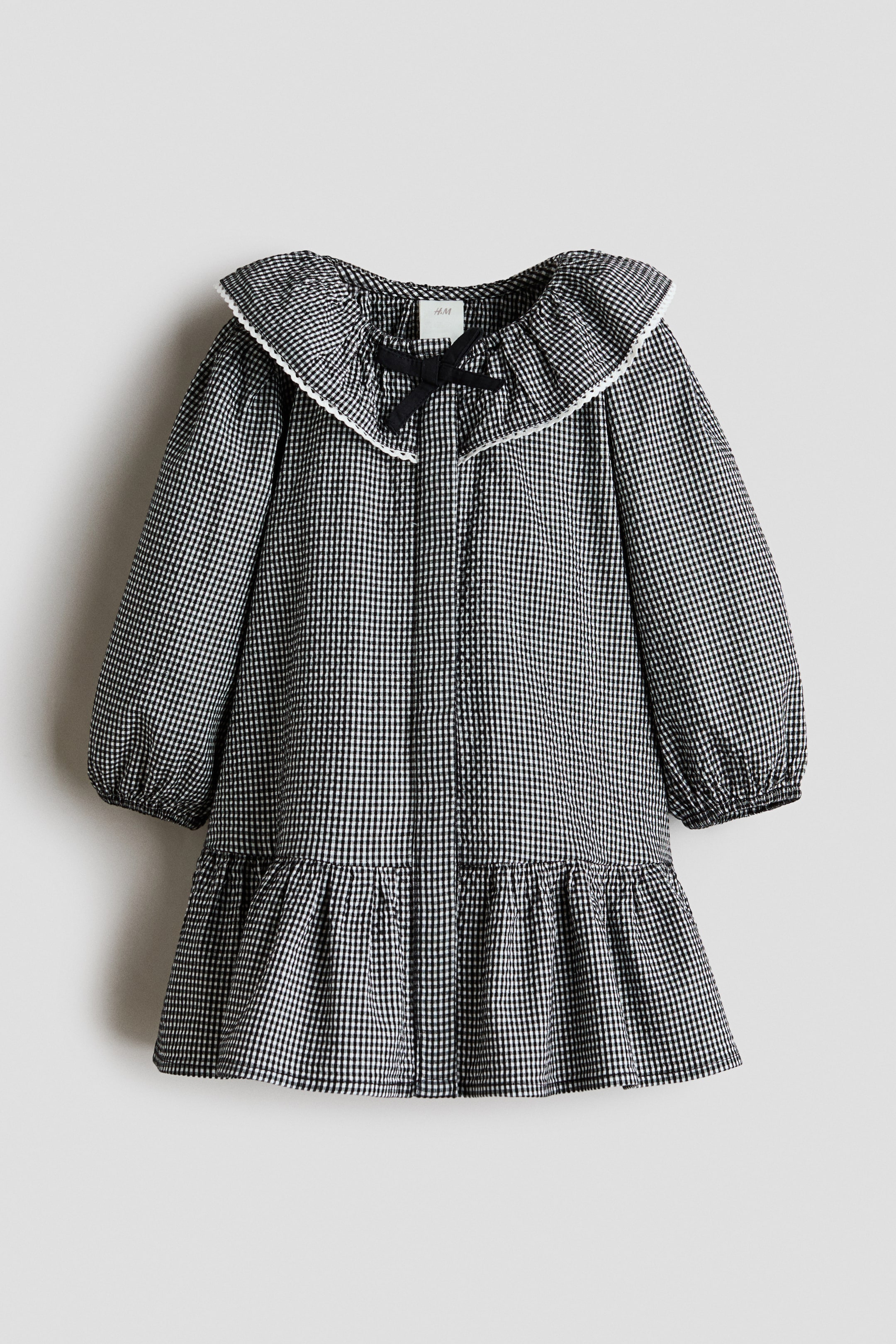 View larger image: A black and white gingham check dress showcasing a large ruffled collar adorned with a white trim and a black bow, complemented by long puffed sleeves and a tiered skirt with a ruffled hem.