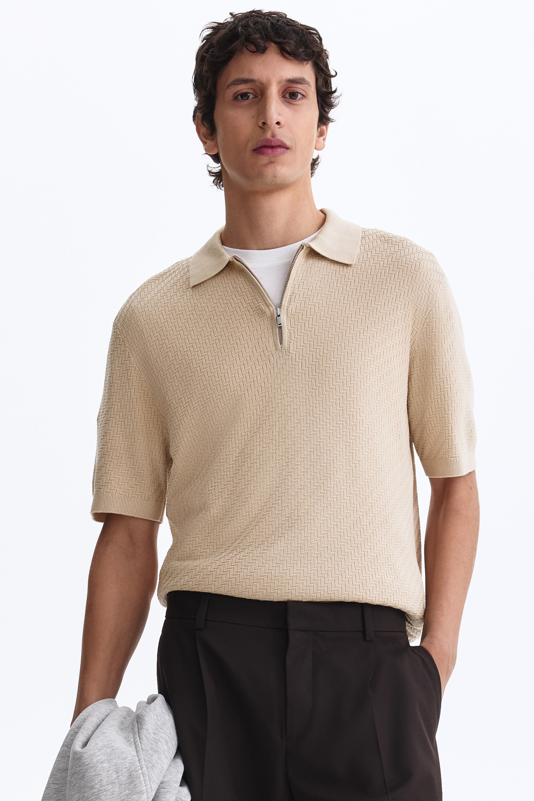 View larger image: A beige knitted polo shirt featuring a herringbone pattern, a classic polo collar, and a quarter-zip opening with a silver-toned zipper. It is designed with short sleeves and ribbed cuffs, and is layered over a white t-shirt.