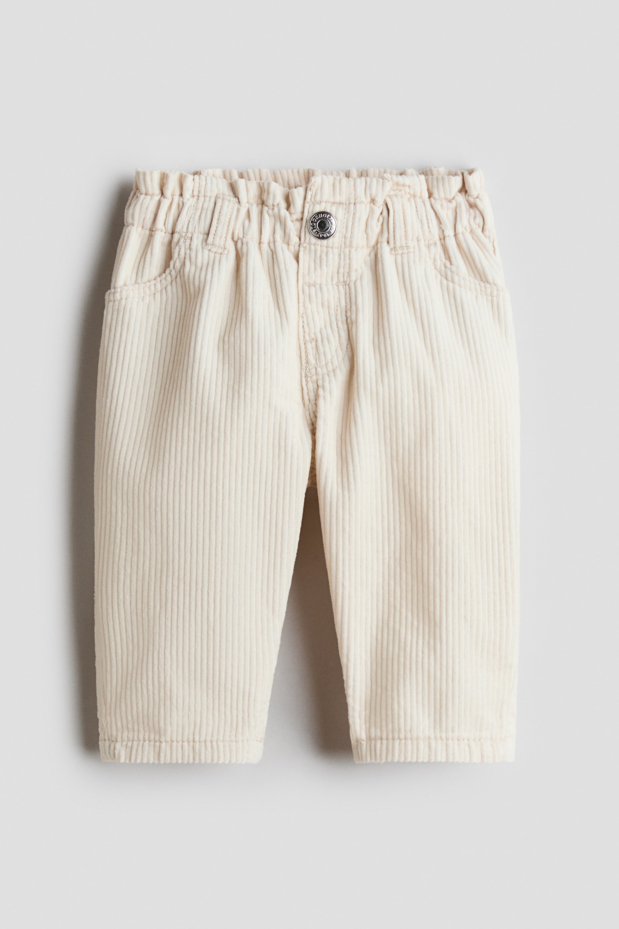 View larger image: Cream corduroy pants are shown facing front, featuring an elasticised waistband with a single metal button closure. They display two angled front pockets and vertical corduroy wales throughout the fabric.
