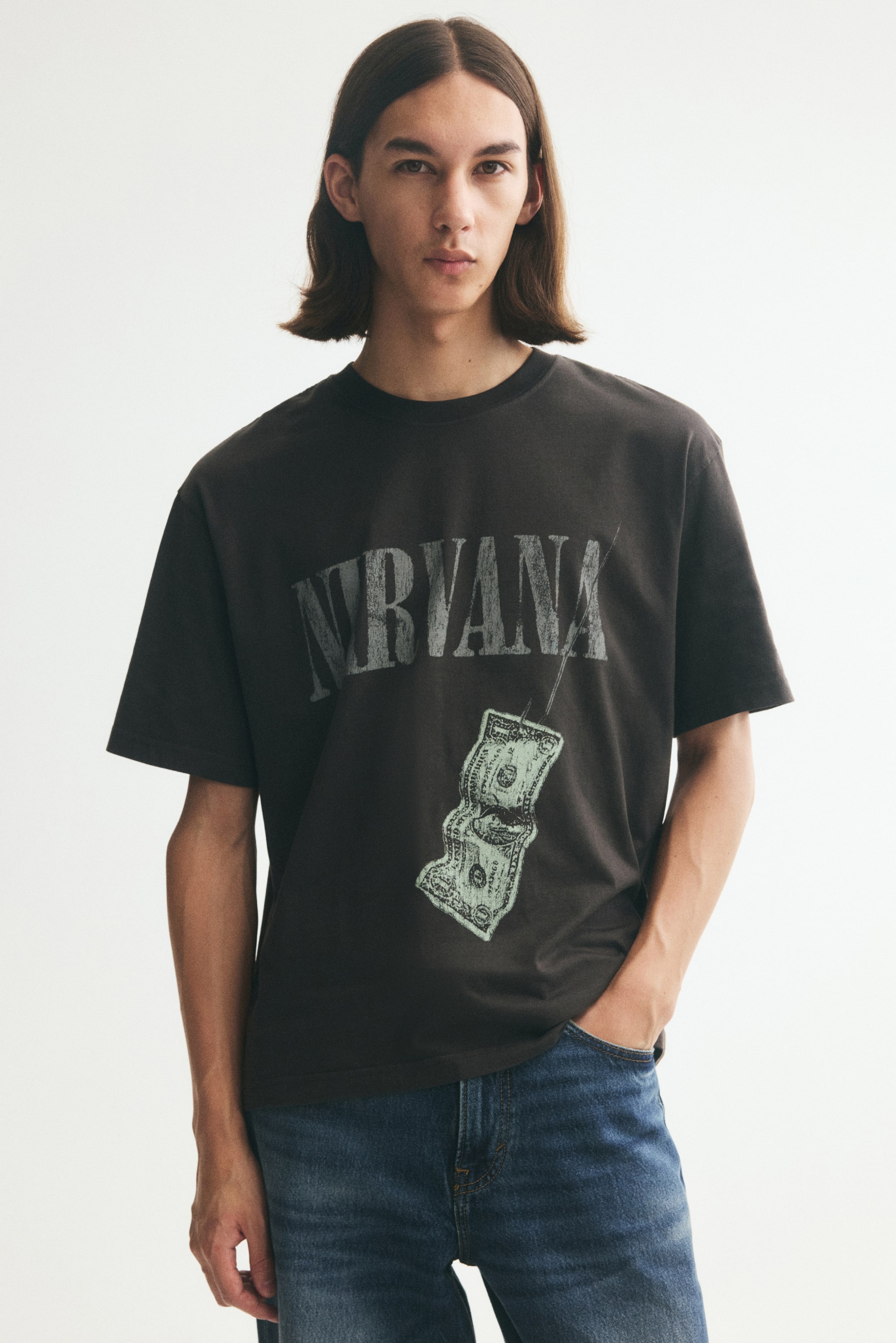 Zobraziť väčší obrázok: Model má na sebe tmavosivé grafické tričko vo voľnejšom strihu s nápisom „Nirvana“ a potlačou dolárovej bankovky. Tričko má okrúhly rebrovaný výstrih a spadnuté ramená a je skombinované s modrými džínsami.