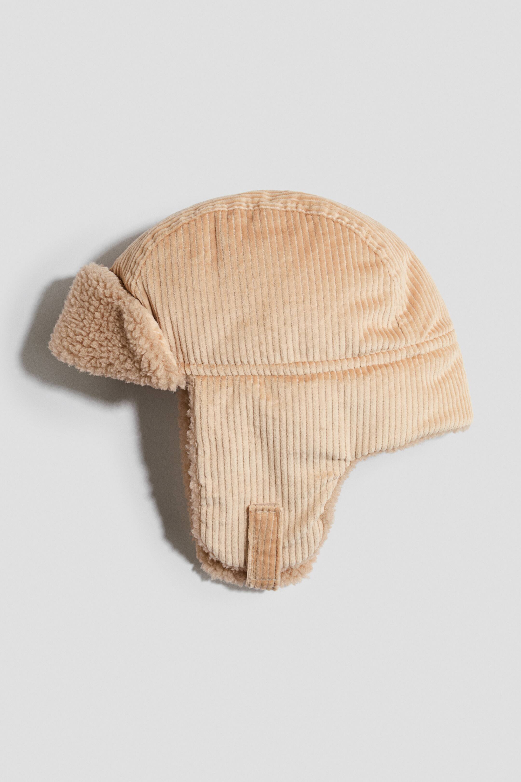 Größeres Bild anzeigen: Eine beigefarbene Cord-Trapper-Mütze liegt flach und zeigt ihr Seitenprofil. Das Design präsentiert eine gerippte Cord-Außenseite, Ohrklappen mit Kinnriemen mit Klettverschluss sowie einen hochgeklappten Frontumschlag, der mit weichem, strukturiertem Teddy-Fleece gefüttert ist.