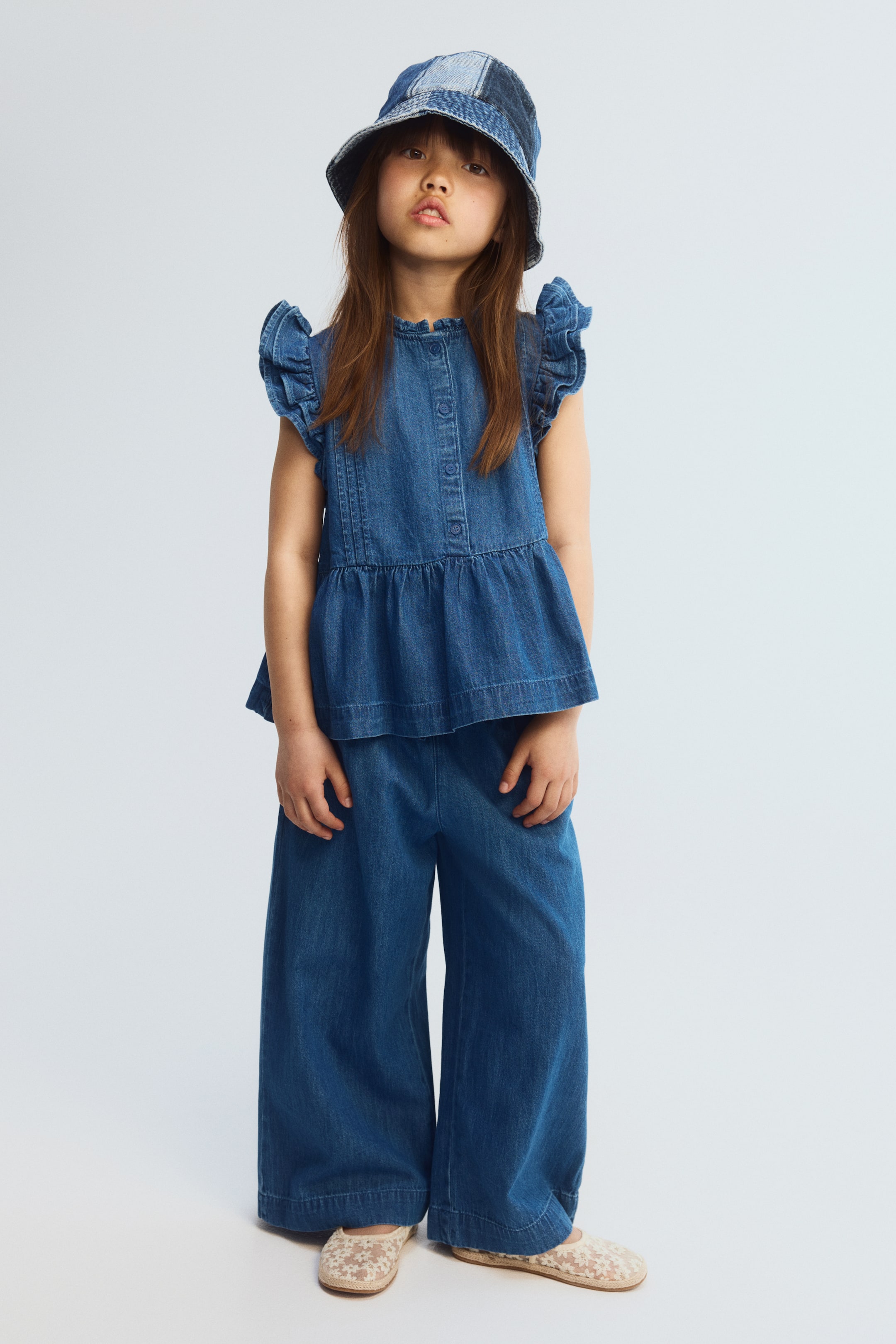 Ampliar la imagen: Se presenta un conjunto de mezclilla para niños, que incluye un sombrero de pescador de mezclilla con diseño de retazos, un top sin mangas con peplum, mangas cortas con volantes y una botonadura frontal, y pantalones de pierna ancha, todo en mezclilla azul y mirando hacia adelante.
