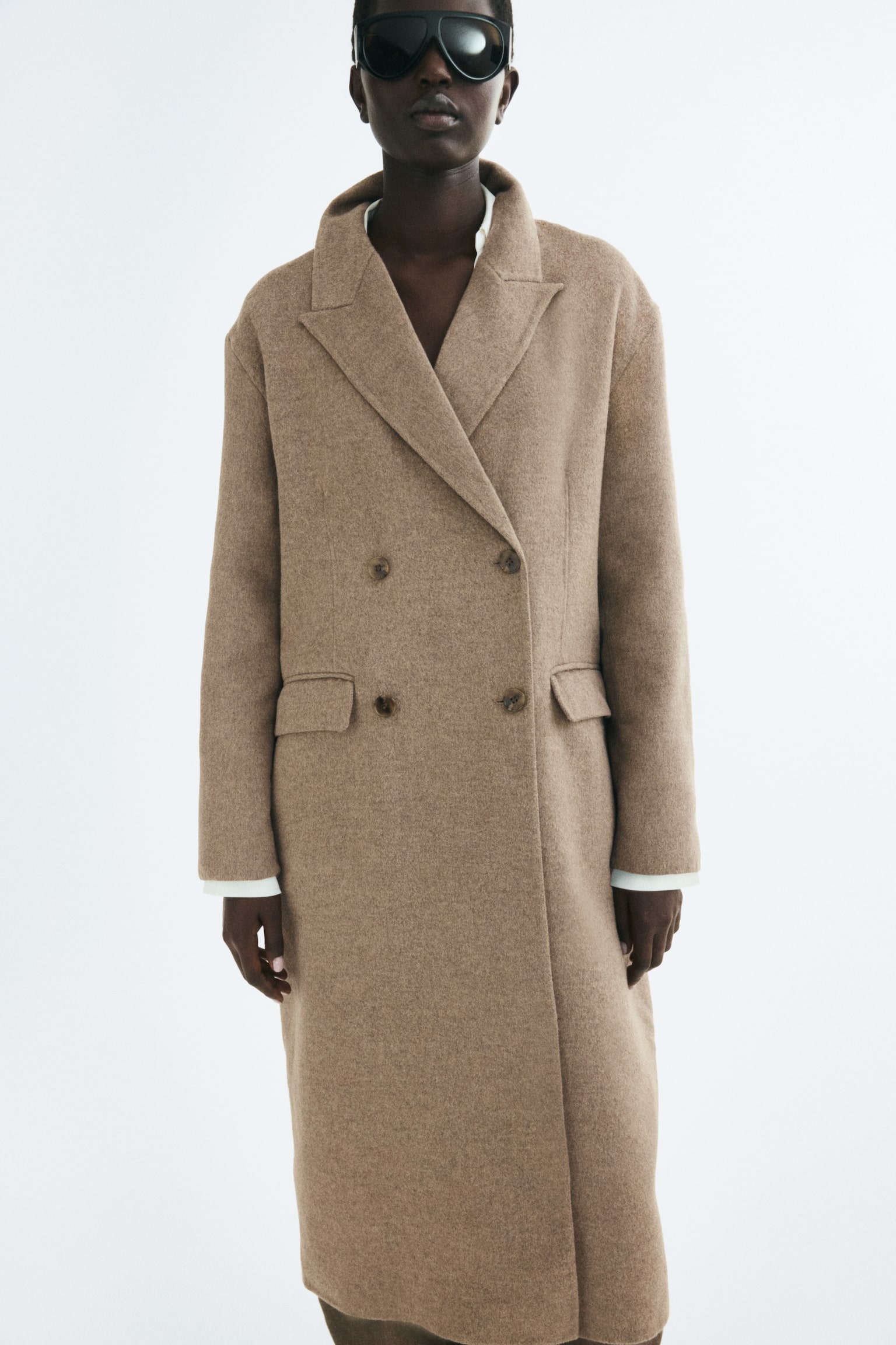 The model is wearing a beige, felted double-breasted maxi coat featuring peak lapels, front button closures, and two side pockets with flaps. The white cuffs of an undergarment are visible at the sleeve openings of the coat. - 1
