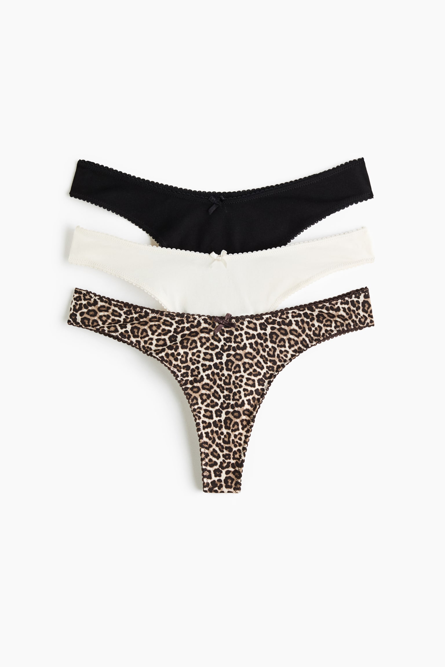 Kolmen puuvillajerseystÃ¤ valmistetun G-string-alushousun pakkaus on kuvattu edestÃ¤, ja siinÃ¤ nÃ¤kyy matalavyÃ¶tÃ¤rÃ¶inen malli, jossa on aaltoileva joustoneulosreunus ja pieni koristeellinen rusetti etukeskellÃ¤. Valikoimaan kuuluu yksi yksivÃ¤rinen musta pari, yksi yksivÃ¤rinen luonnonvalkoinen pari ja yksi leopardikuosinen pari pinottuna pystysuoraan.