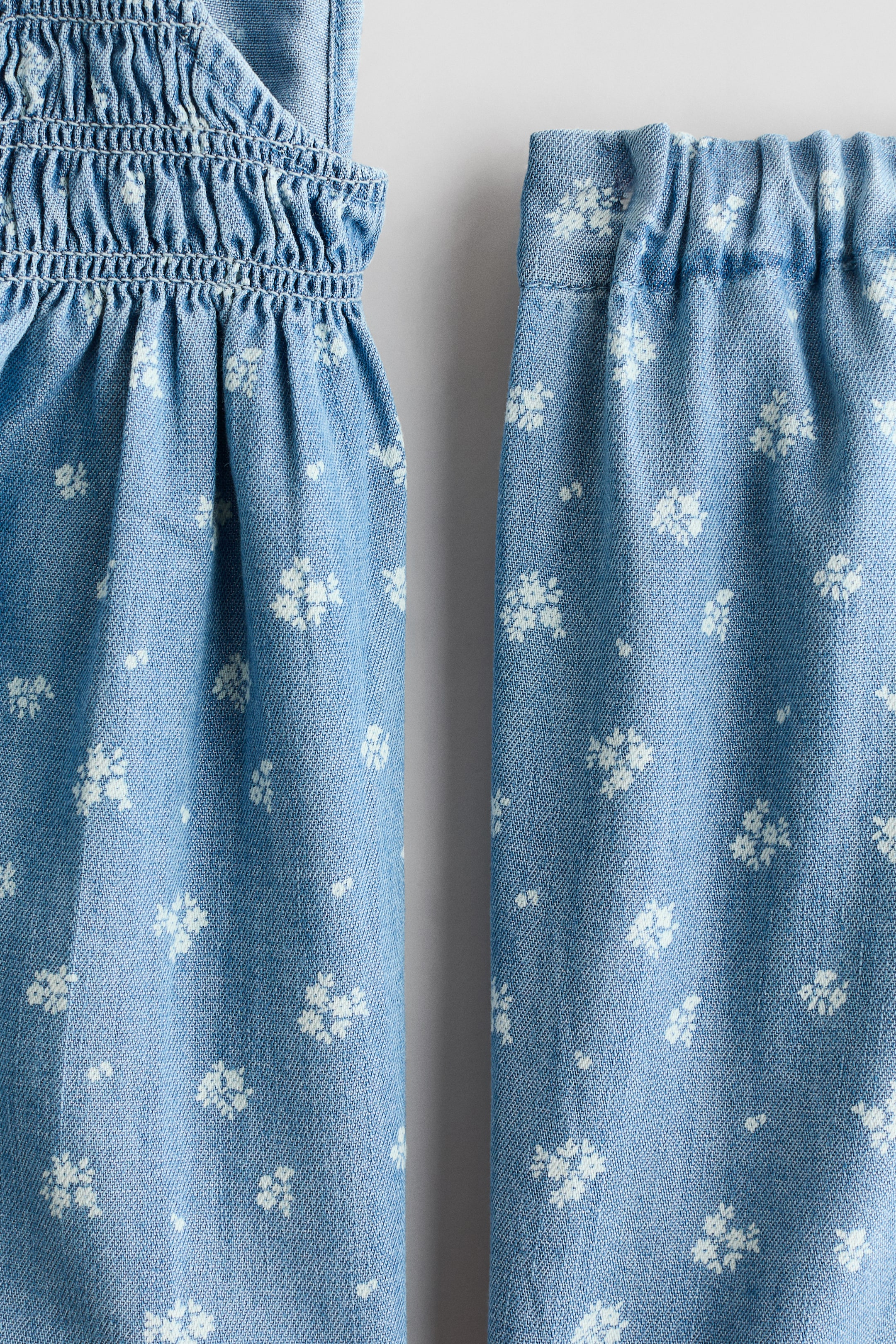 View larger image: A light blue outfit set features a white floral pattern on both pieces. The left piece displays a smocked panel above gathered fabric, while the right piece shows an elasticized waistband leading into gathered fabric.