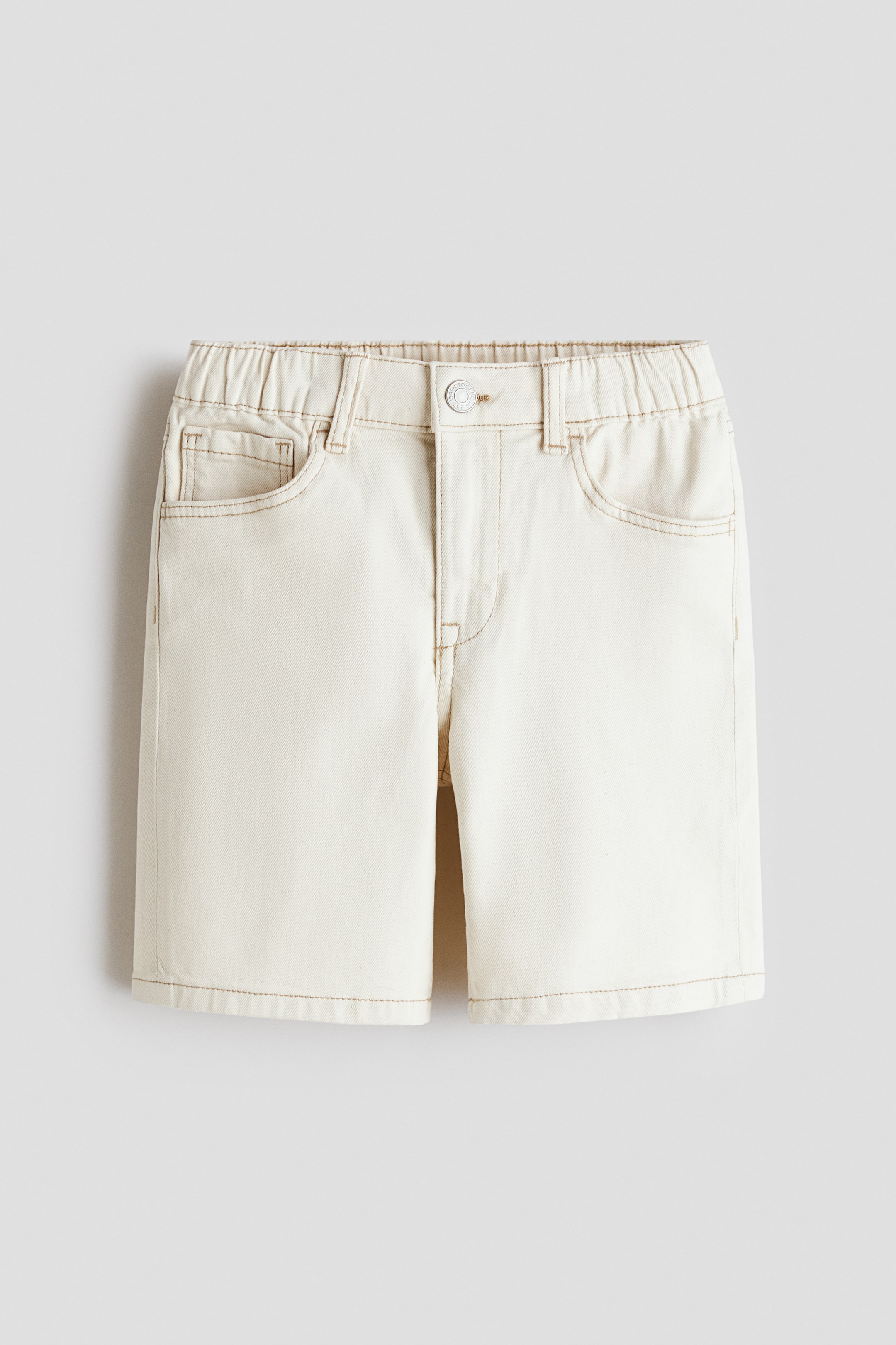 View larger image: Cream shorts are lying flat, featuring an elasticated waistband with a single silver button fastening, two rounded front pockets, a small coin pocket, and contrasting tan stitching throughout.