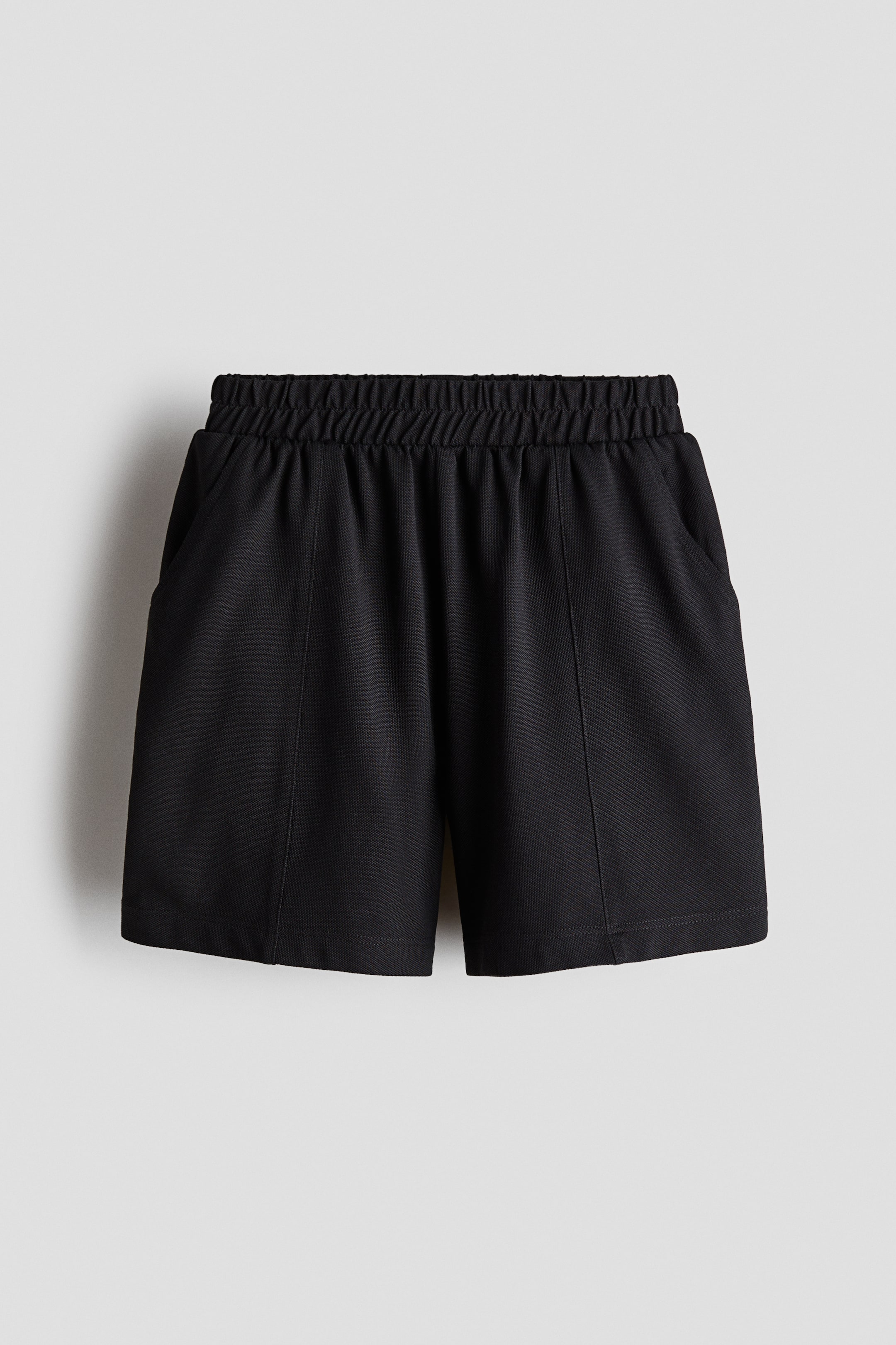 Xem ảnh lớn hơn: Một chiếc quần short màu đen được chụp từ phía trước, làm nổi bật phần cạp chun nhún và túi chéo hai bên. Quần short có các đường may dọc chạy xuống mặt trước của mỗi ống quần rộng, mang đến phom dáng vừa có cấu trúc vừa thoải mái.