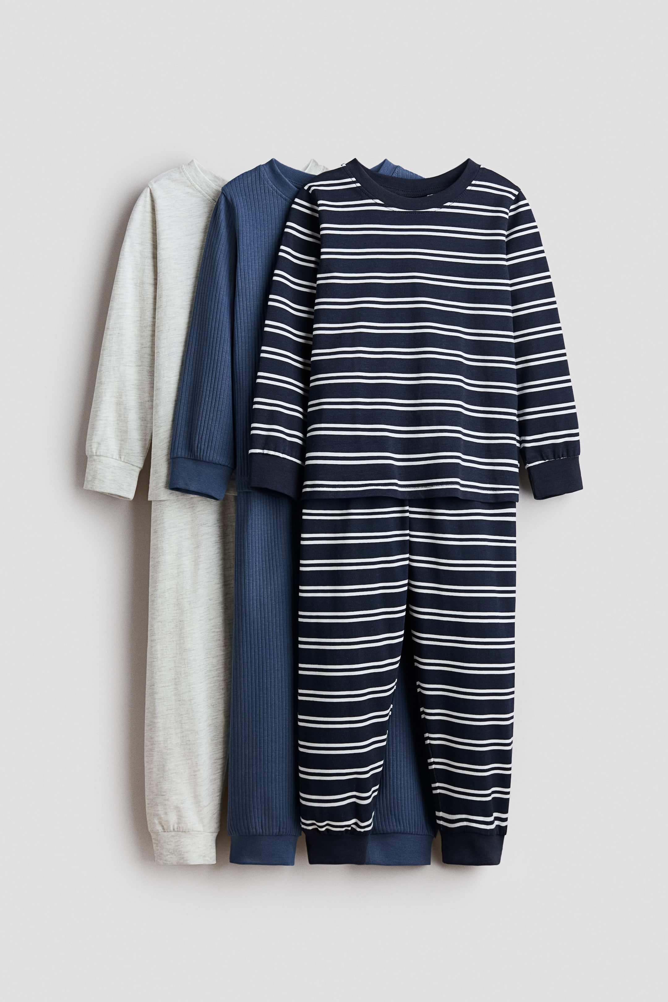 Größeres Bild ansehen: Drei Sets langärmeliger Pyjamas mit langen Hosen hängen vertikal und nach vorne gerichtet. Sie bestehen aus einem schlichten hellgrauen Set, einem dunkelblauen gerippten Set und einem marineblauen Set mit weißen horizontalen Streifen.