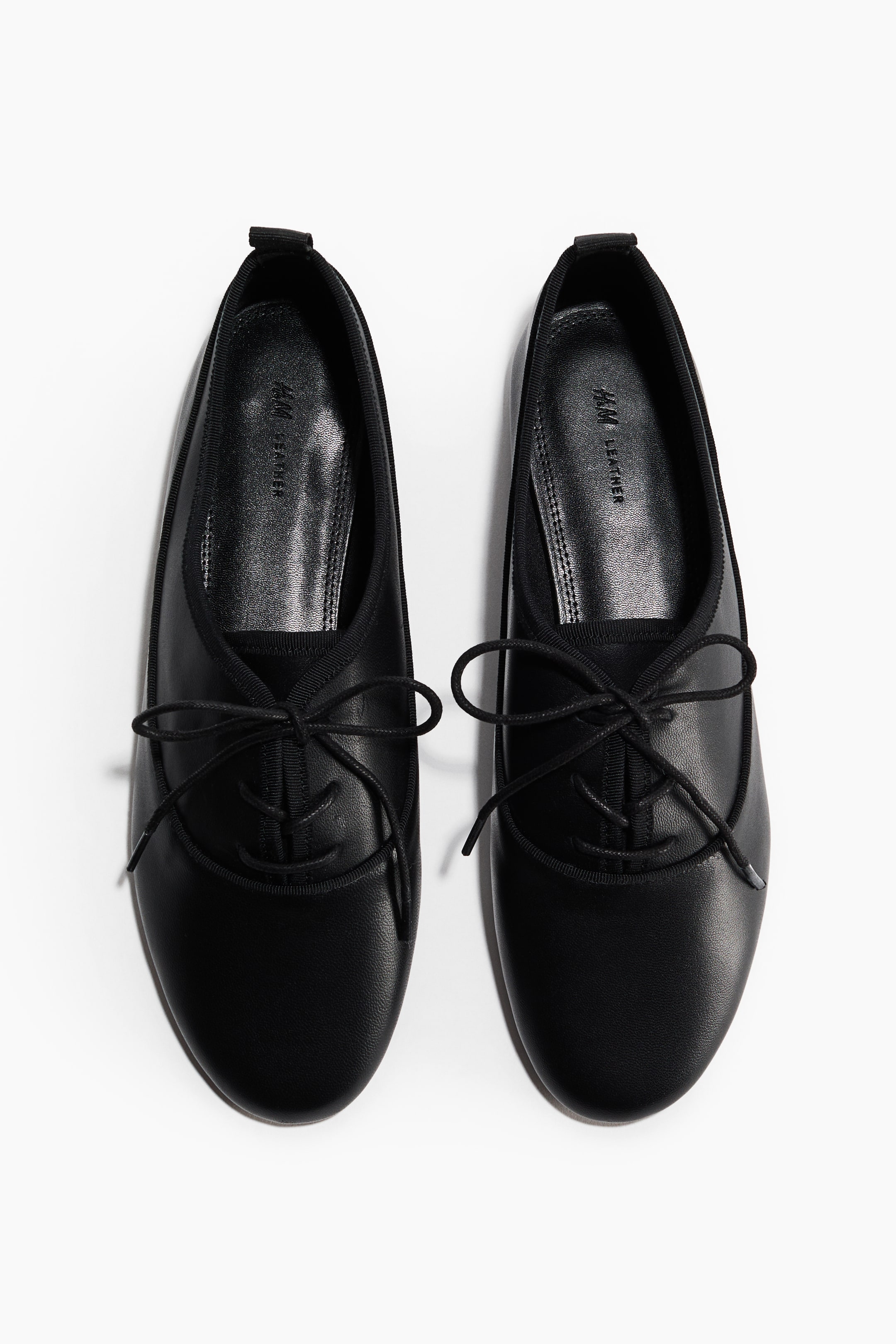 View larger image: A pair of black leather flat shoes is angled slightly inwards, showcasing rounded toes and elasticized openings. Black laces are tied in bows over the vamp of each shoe, and "H&M LEATHER" is visible on the insole.