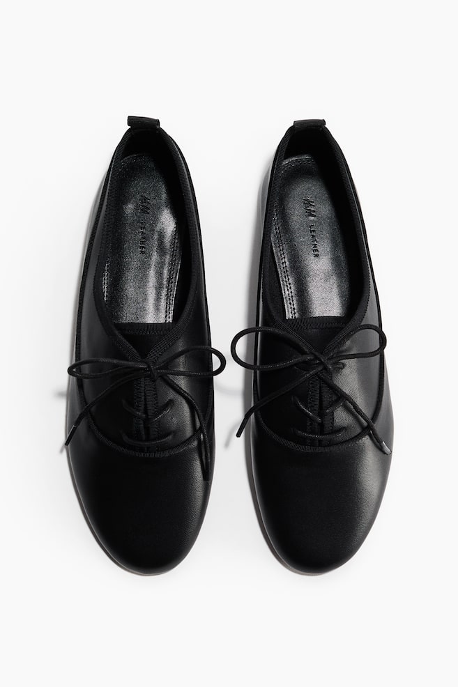 Un par de zapatos planos de cuero negro está ligeramente inclinado hacia adentro, mostrando puntas redondeadas y aberturas elásticas. Los cordones negros están atados en la parte superior de cada zapato, formando lazos sobre el empeine, y "H&M LEATHER" es visible en la plantilla. - 1